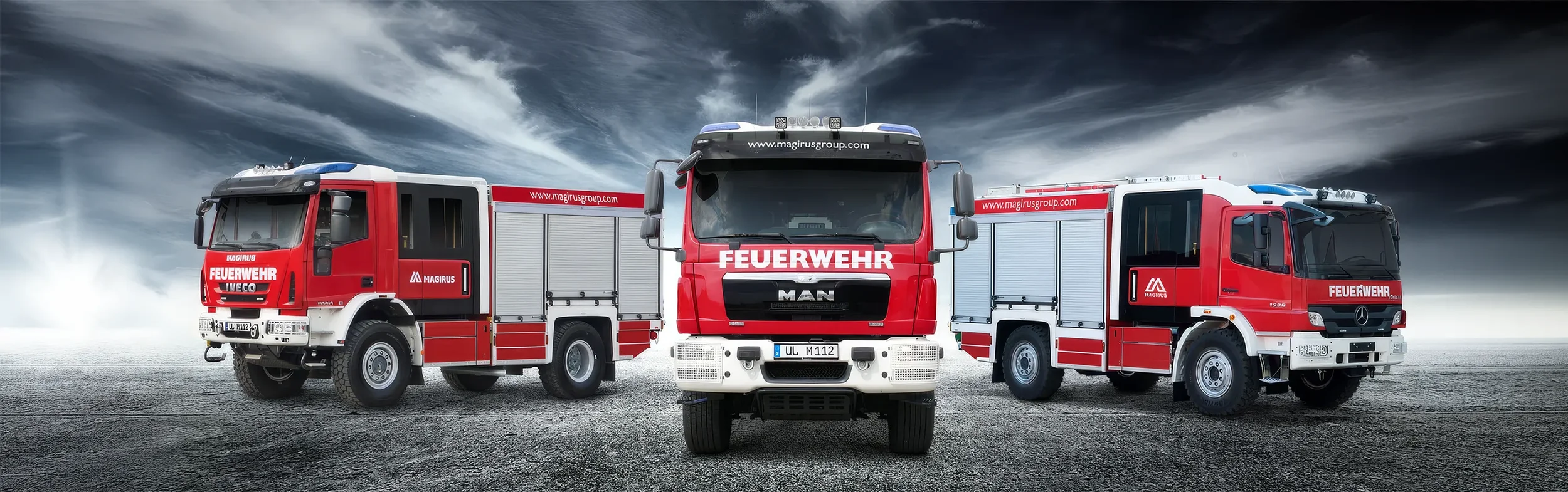 Three red and white fire trucks are parked on a gravel surface under a cloudy sky. The trucks are labeled with German words for fire department and wear the insignia of firefighting units.