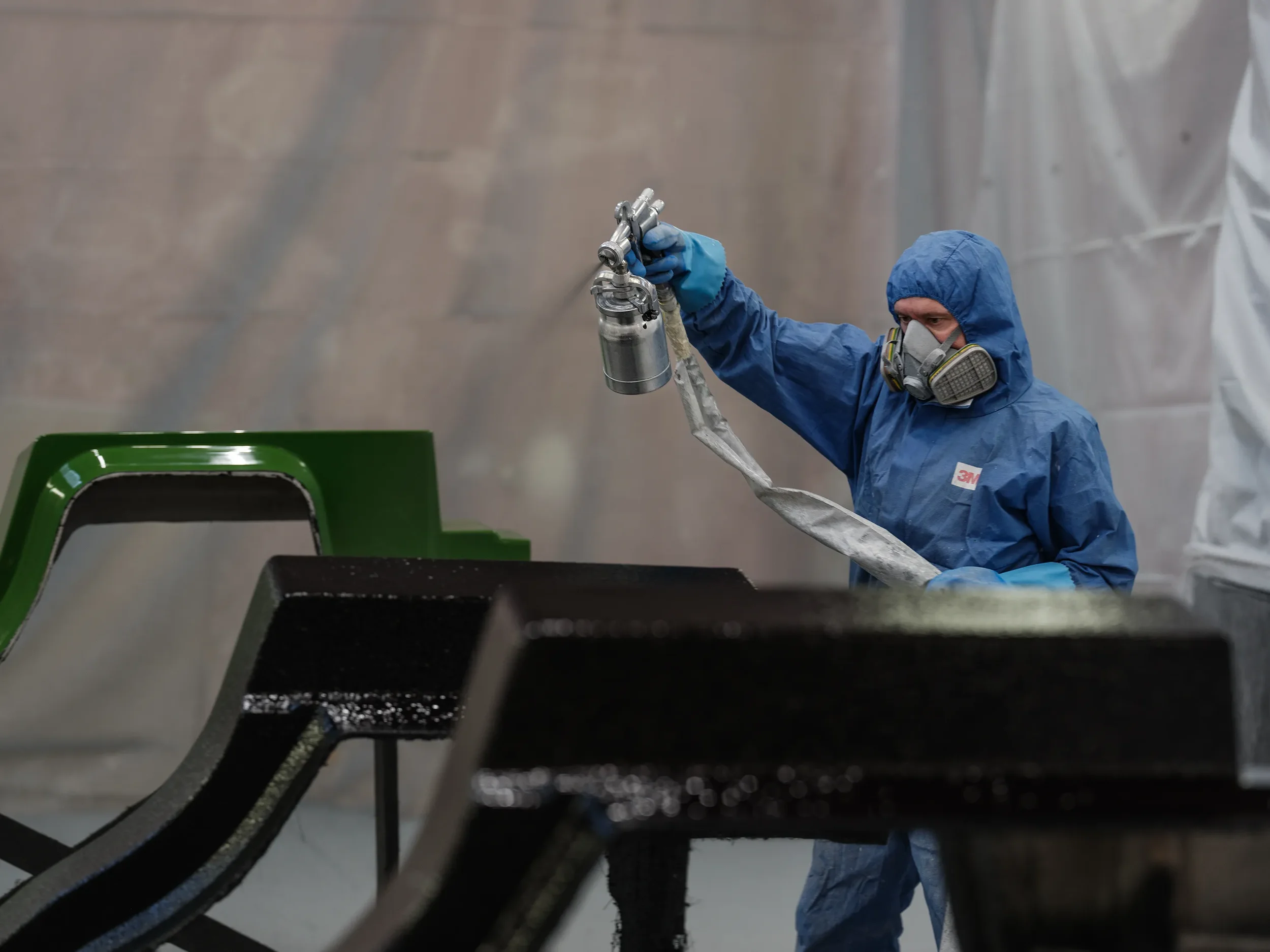 A worker in blue protective gear and a respirator mask is spray painting a car bumper with a spray gun in a painting facility.