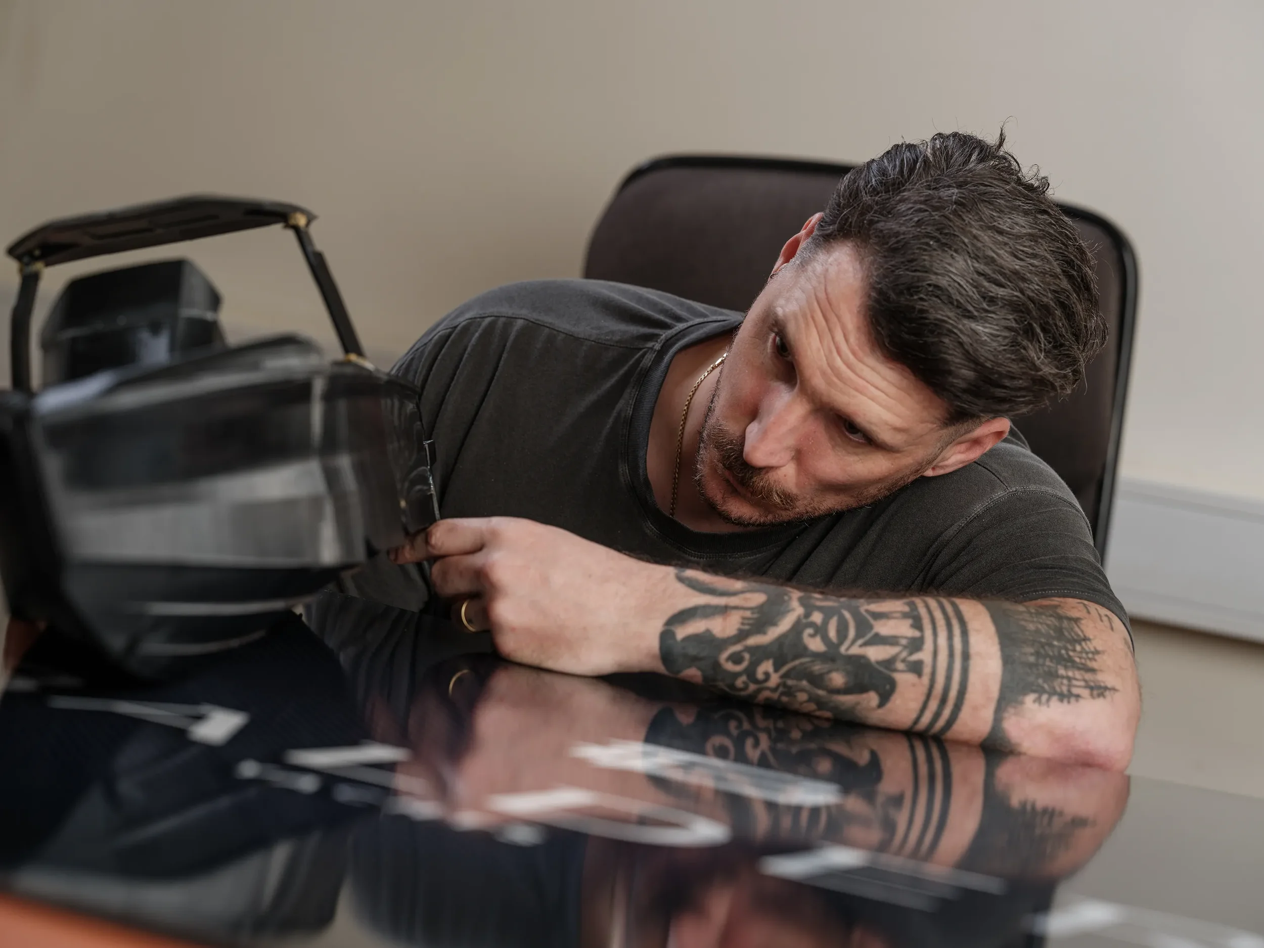 Man with tattoos on arm opening a black briefcase on a reflective table in an office setting.