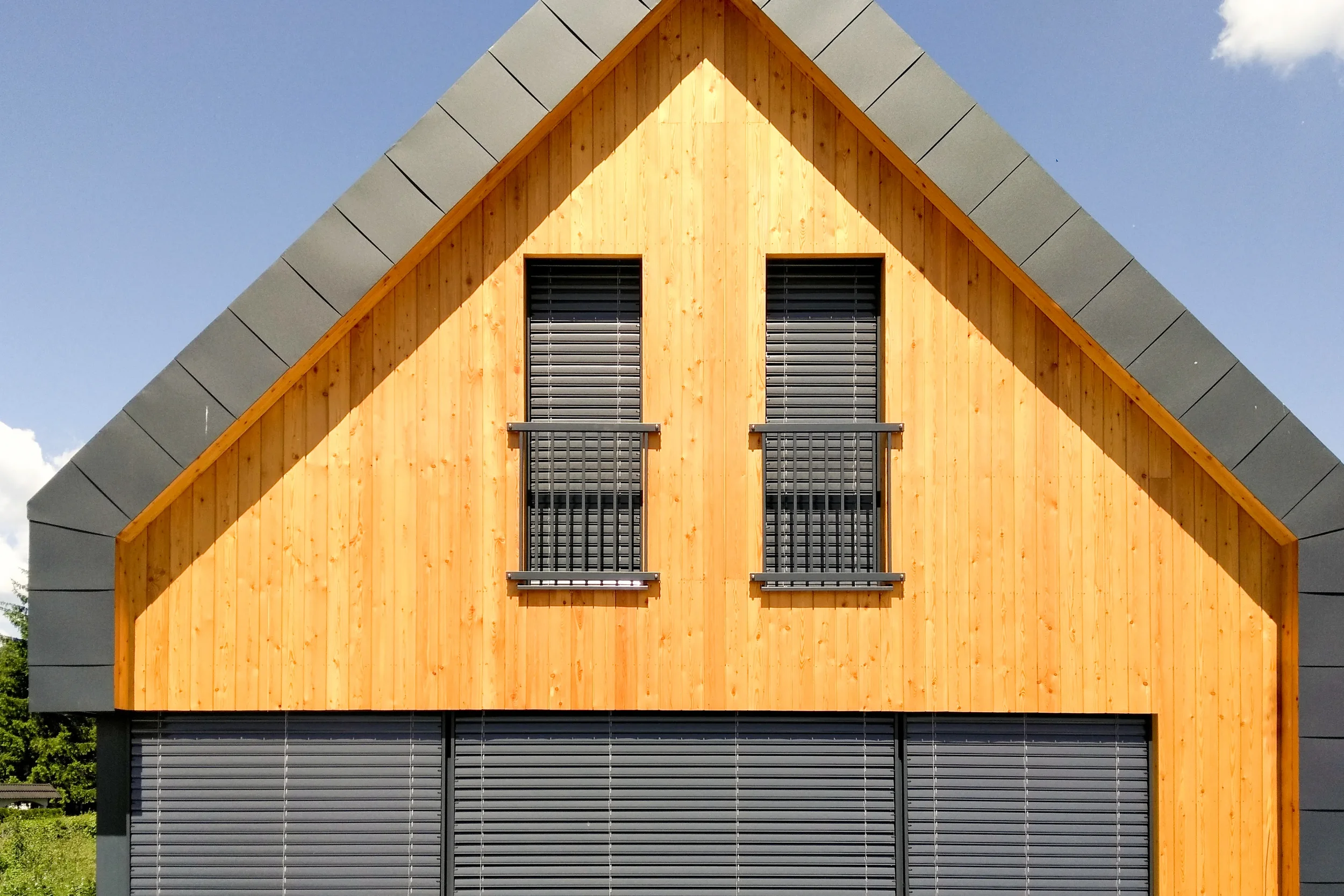Close-up of a modern house with a wooden facade, gray metal window shutters, and a gray metal roof against a blue sky.