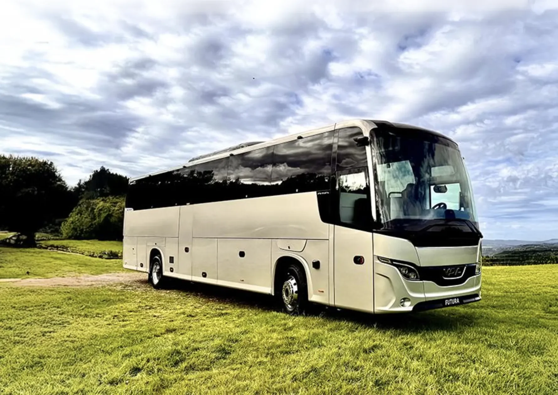 A white and black tour bus parked on a grassy field with trees and cloudy sky in the background.