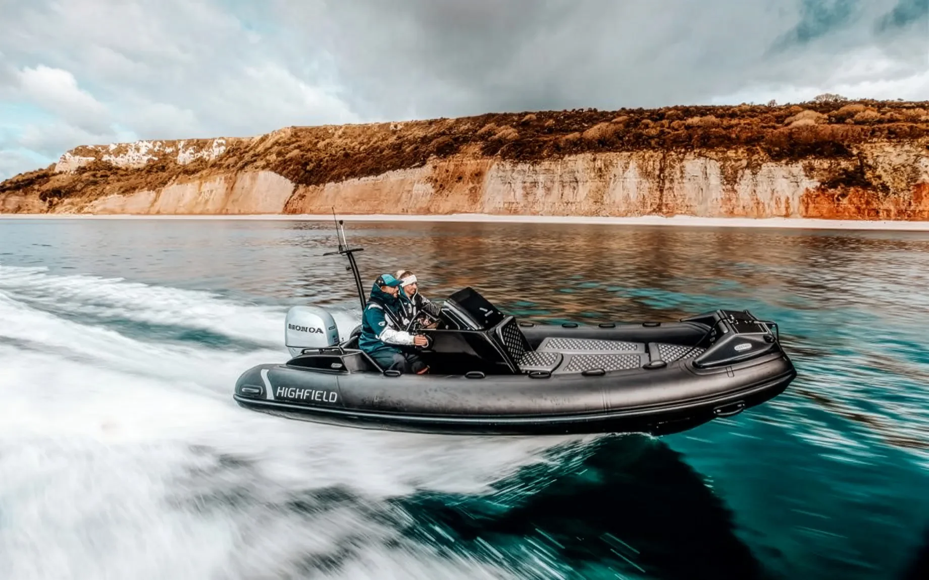 Two people on a black Highfield rigid inflatable boat speeding on water near a rocky coastline with cliffs.