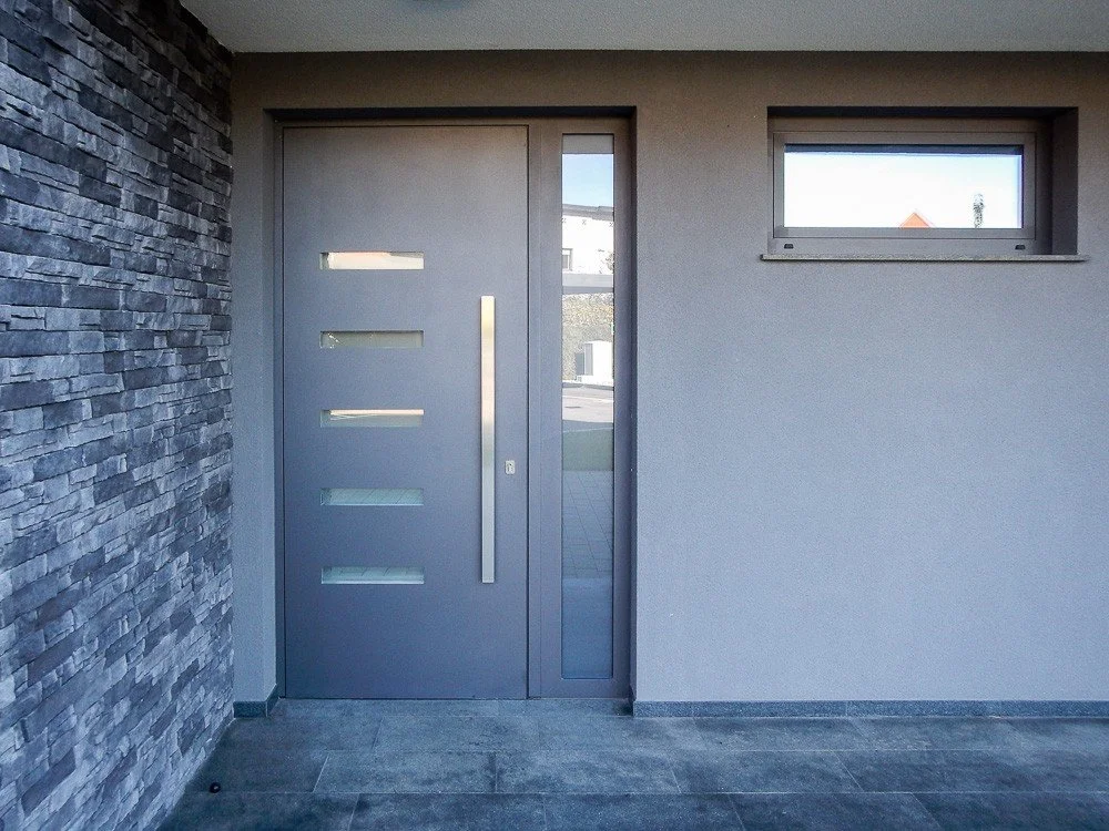 Modern house entrance with a gray door featuring horizontal glass panels, side glass panel, stone wall on the left, and small rectangular window on the right wall.