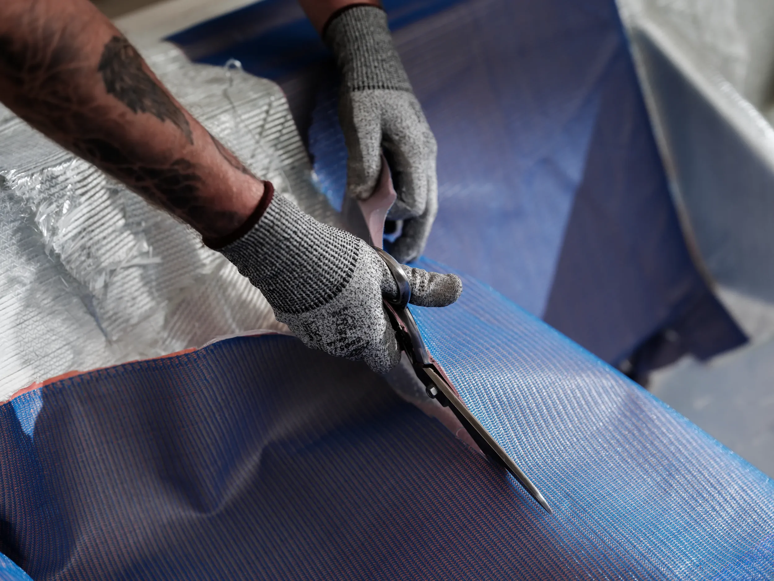 Close-up of a worker's hands cutting a blue, textured fabric with scissors, wearing gray gloves with black accents in an industrial setting.