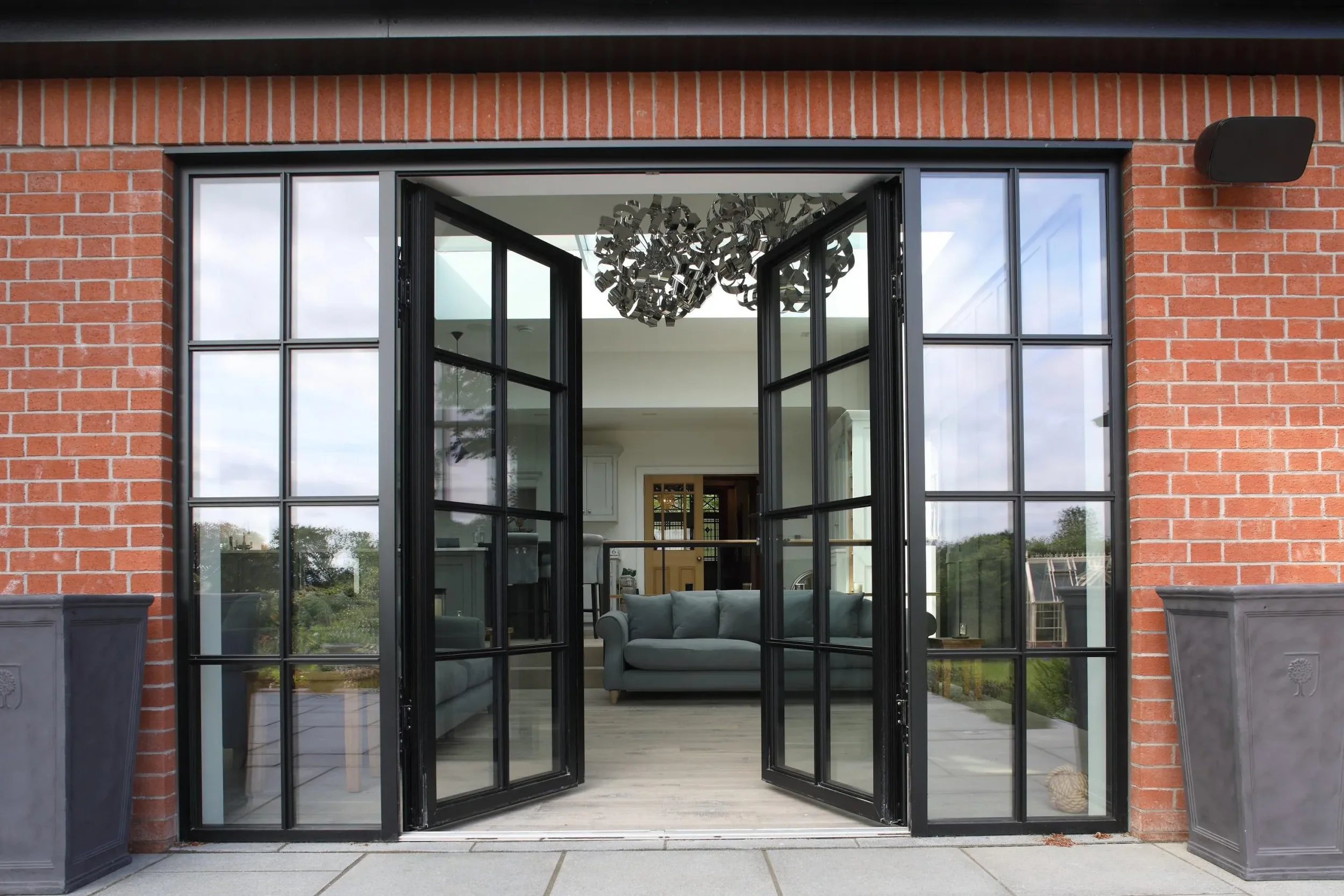 Open black-framed glass door leading to a modern living room with gray sofas and a contemporary chandelier, brick exterior wall.