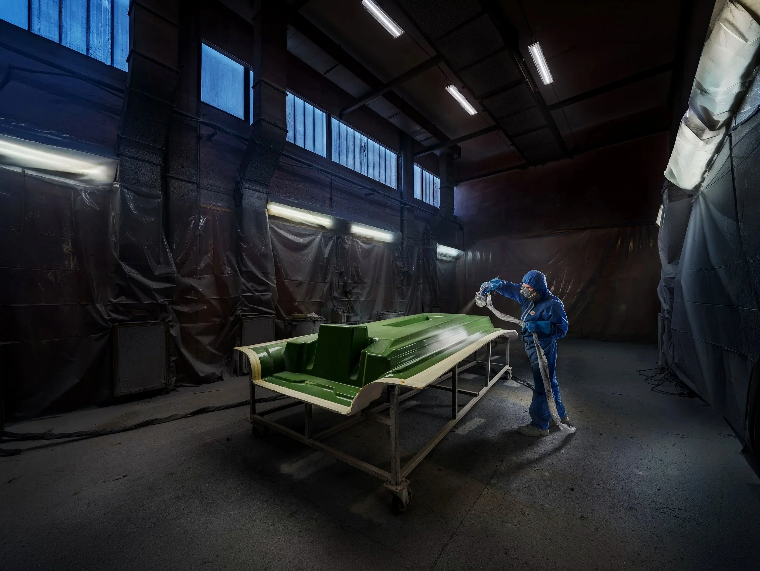 A person in blue protective gear spray-paints or coats a moss-green kayak inside a workshop with black walls, high windows, and ceiling lights.