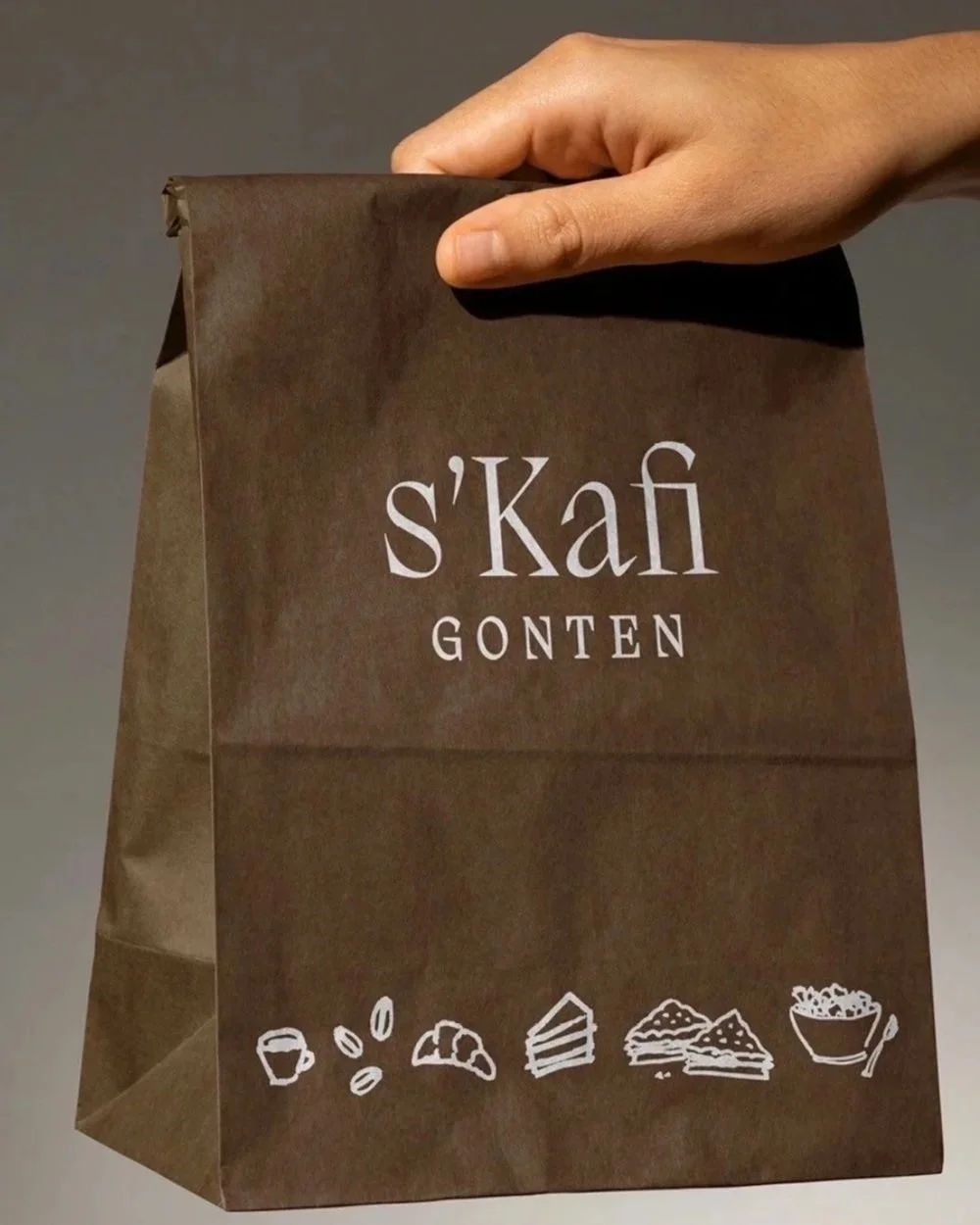 Eine braune Papiertüte mit weissen Symbolen für Kaffee, Nüsse, Croissants, Kuchen, Kekse und Müsli, sowie dem Text 's'Kafi GONTEN' auf der Vorderseite.