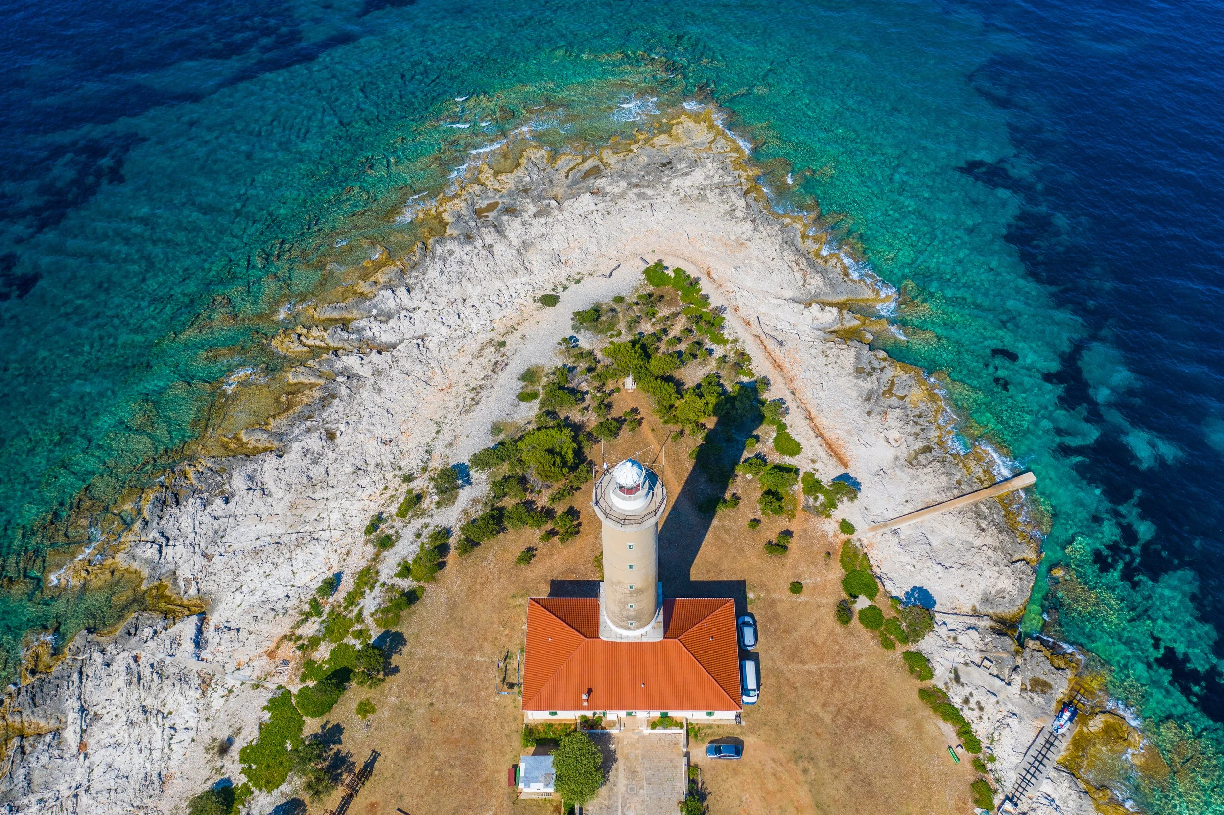 Aerial view of a lighthouse on a rocky coastline with trees and a red-roofed building nearby, surrounded by clear blue ocean water.