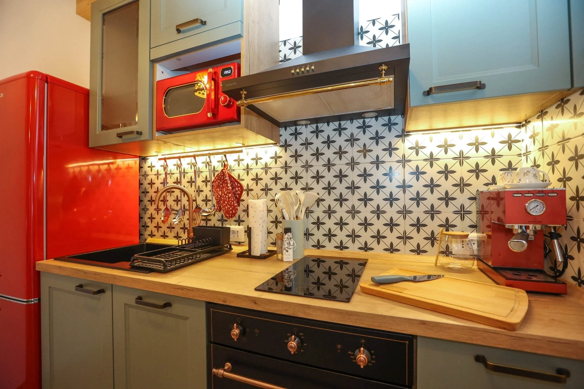 A kitchen featuring a red retro refrigerator, a black stove with copper knobs, a wooden countertop with utensils, a red espresso machine, and patterned black and white tile backsplash with floral design. Cabinets are painted light blue and gray.