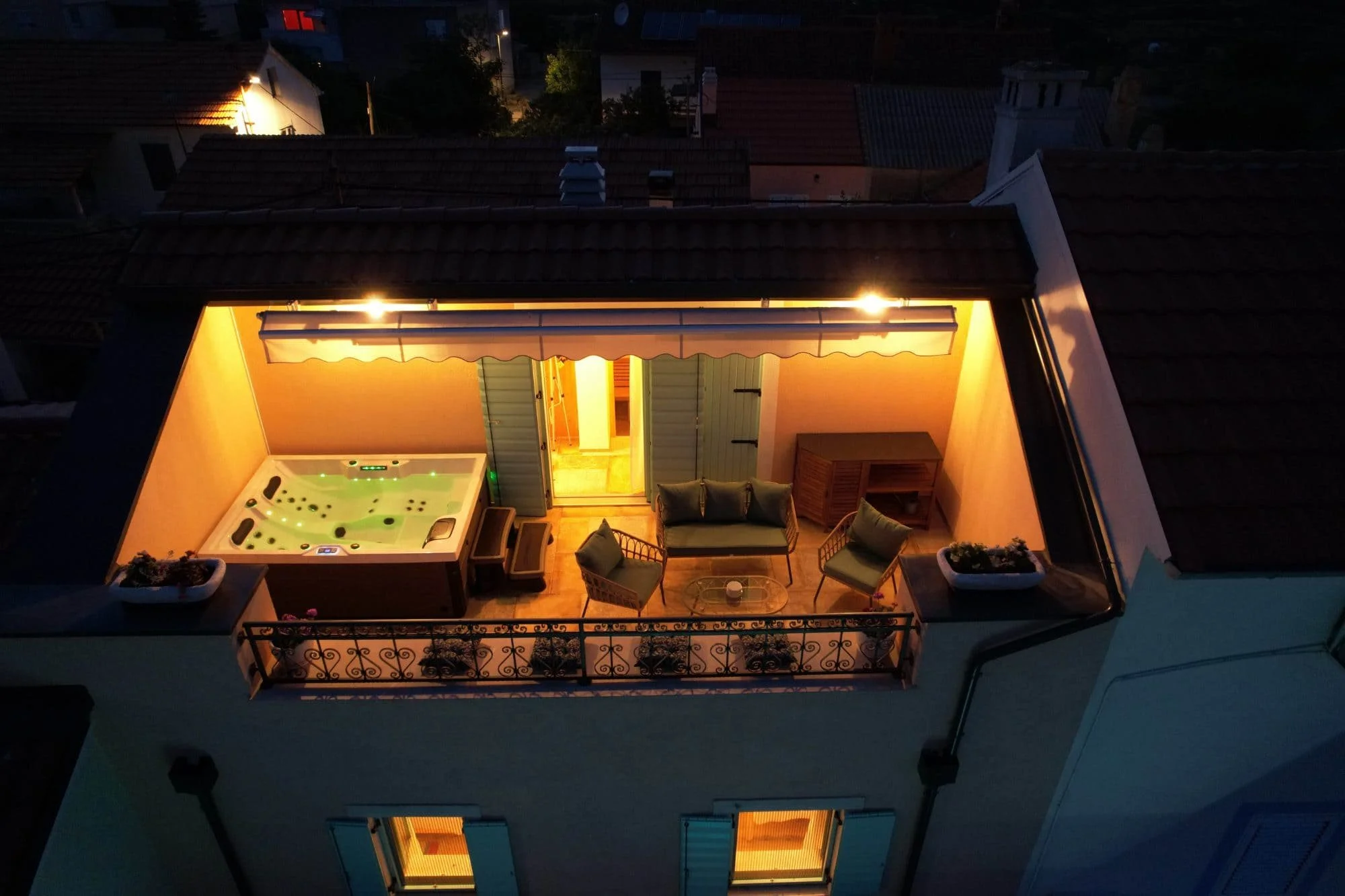 A rooftop balcony at night with warm lighting, furnished with outdoor chairs, a sofa, and a glass table, overlooking neighboring rooftops and houses in a residential area.