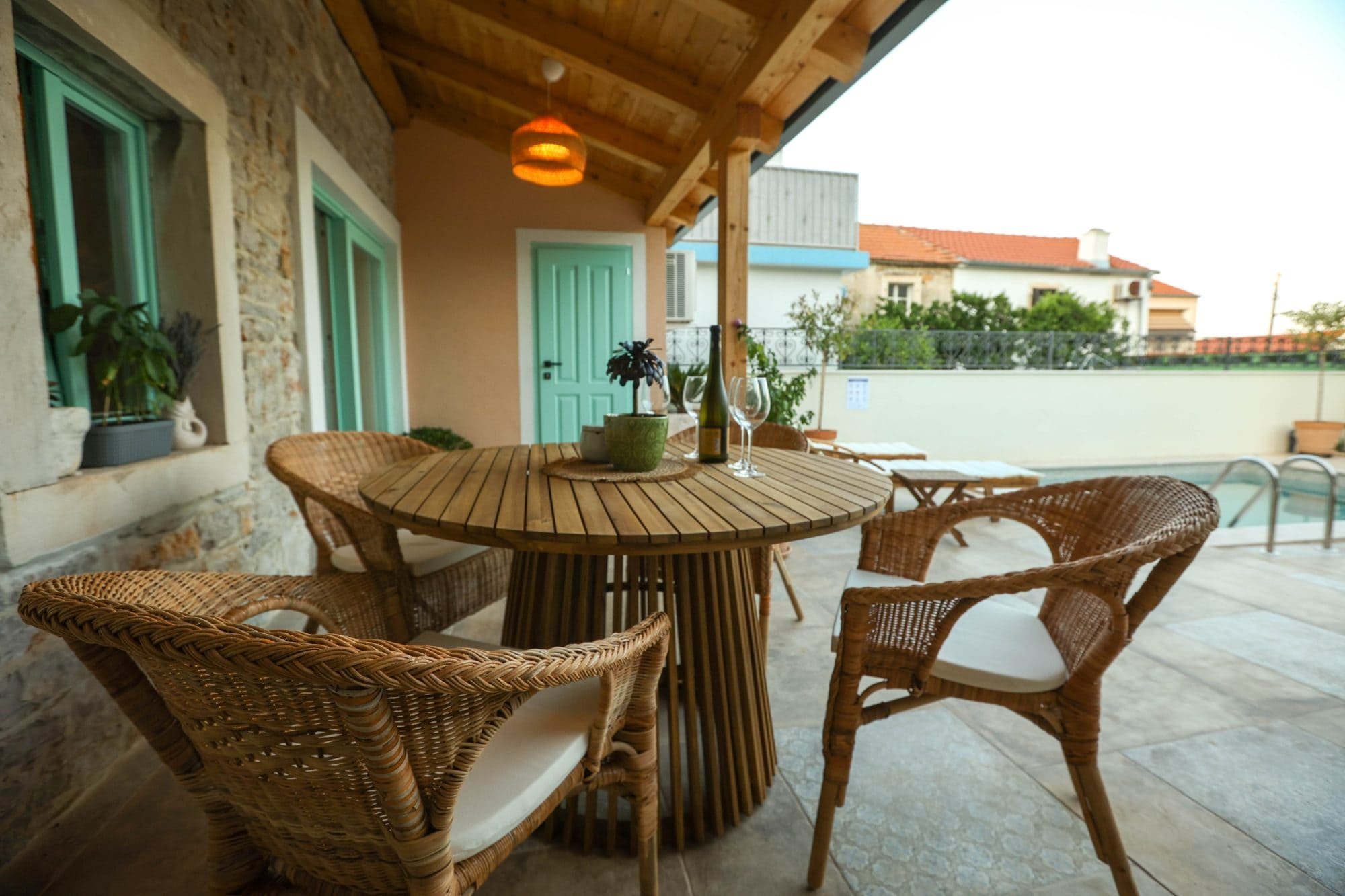 Outdoor patio area with a round wooden table, wicker chairs, and wine glasses, with a house wall featuring windows, a door, and a wooden ceiling overhead.