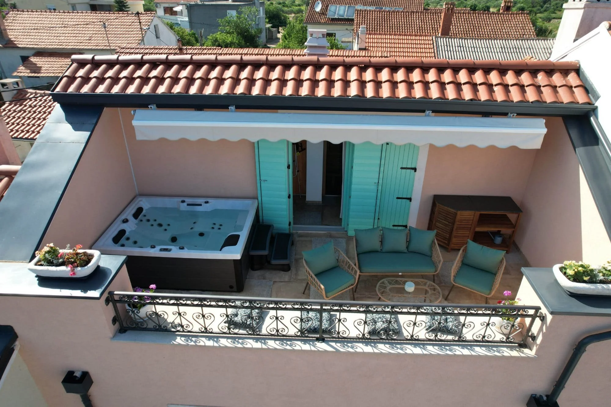 A rooftop terrace with a hot tub, outdoor furniture including a couch and chairs, potted plants, and a retractable awning, overlooking neighboring houses with red tile roofs and green trees.