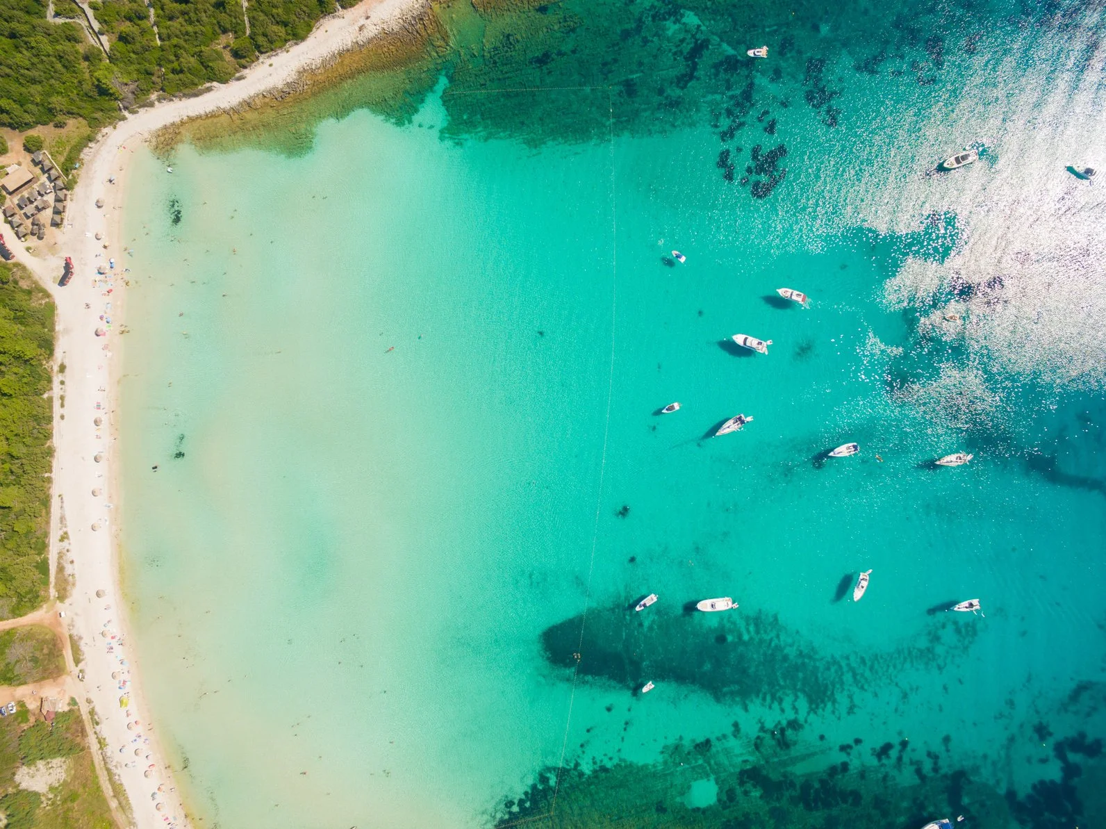 An aerial view of a beach with turquoise waters, a sandy shore, and boats anchored in the water. Green trees line the beach on the left side.