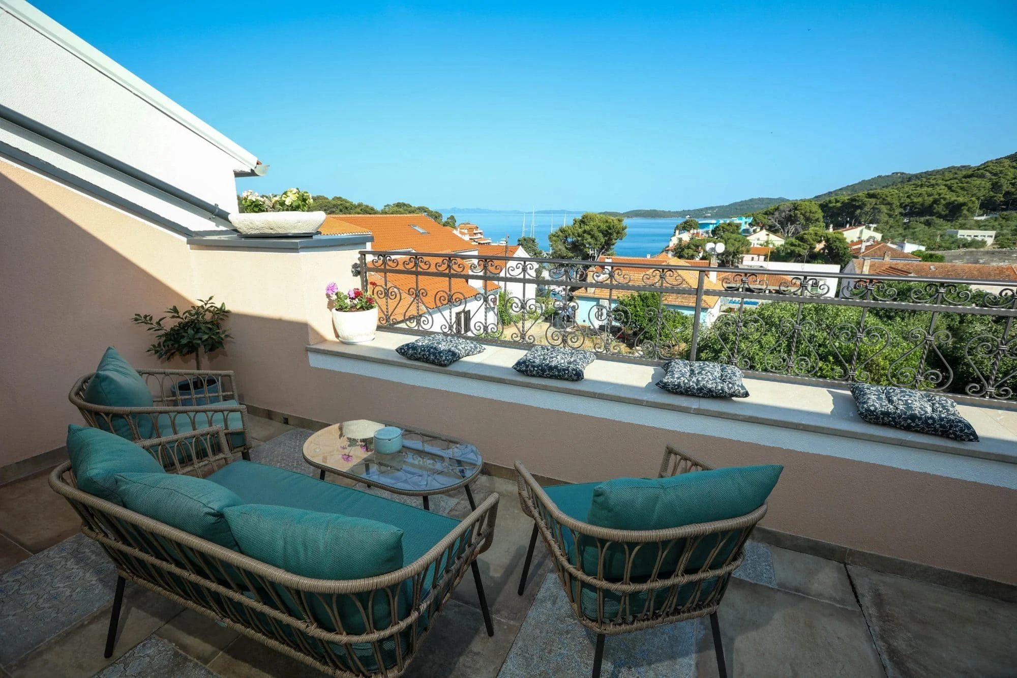 A cozy balcony with outdoor seating including couches and chairs with teal cushions, a small round table, potted plants, and decorative cushions. The balcony offers a view of rooftops, green trees, and a body of water with sailboats, hills, and a cle