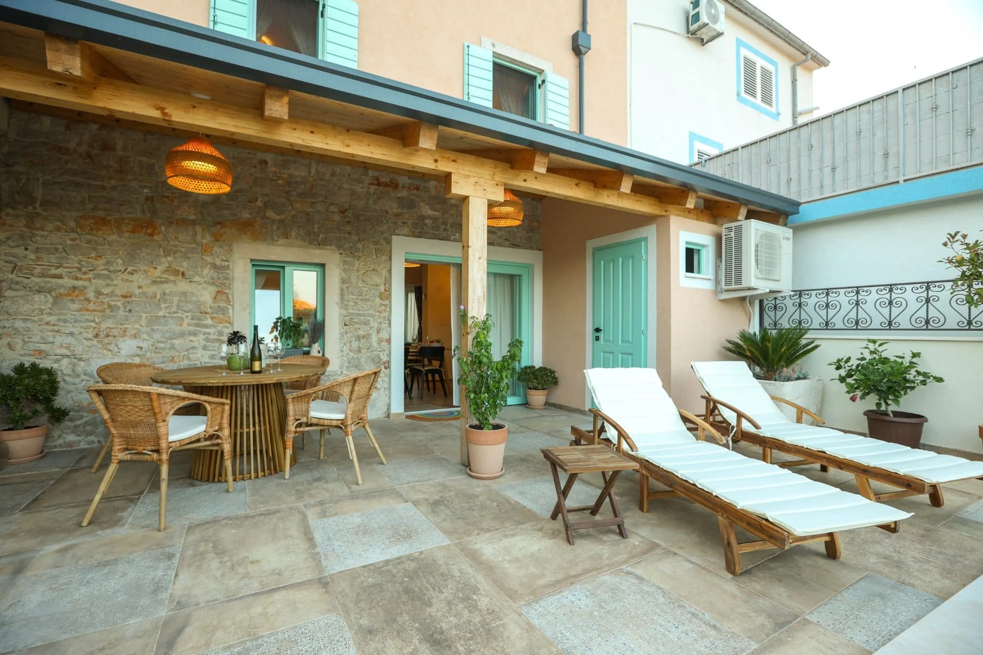 A cozy outdoor patio area with a stone wall, wooden ceiling, and hanging wicker lights. There is a round wooden dining table with wicker chairs, some potted plants, lounge chairs with white cushions, and a small wooden side table. The backdrop includ