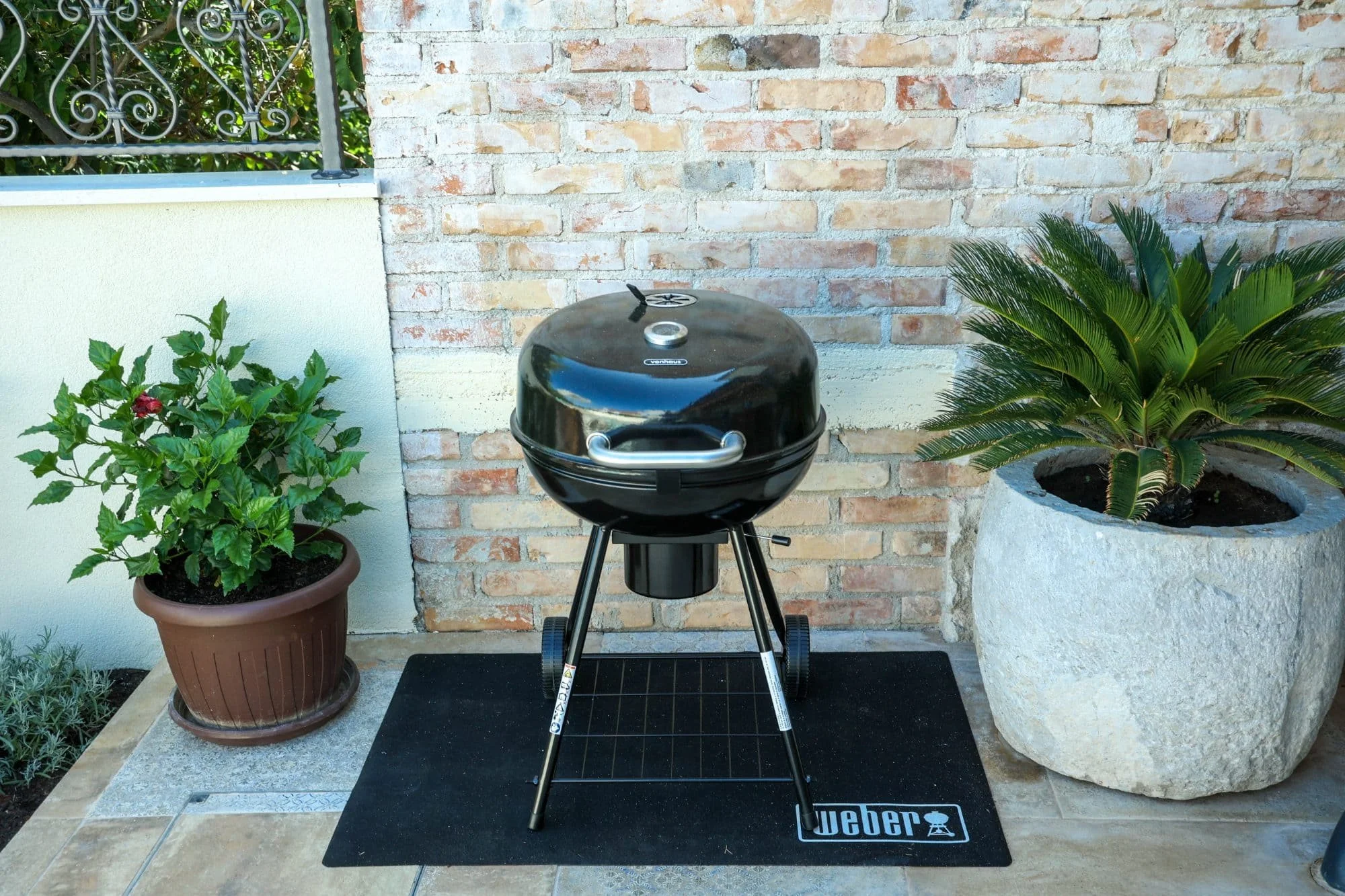 A black Weber charcoal grill on a patio with two potted plants on either side, a brick wall in the background, and a black Weber mat underneath.