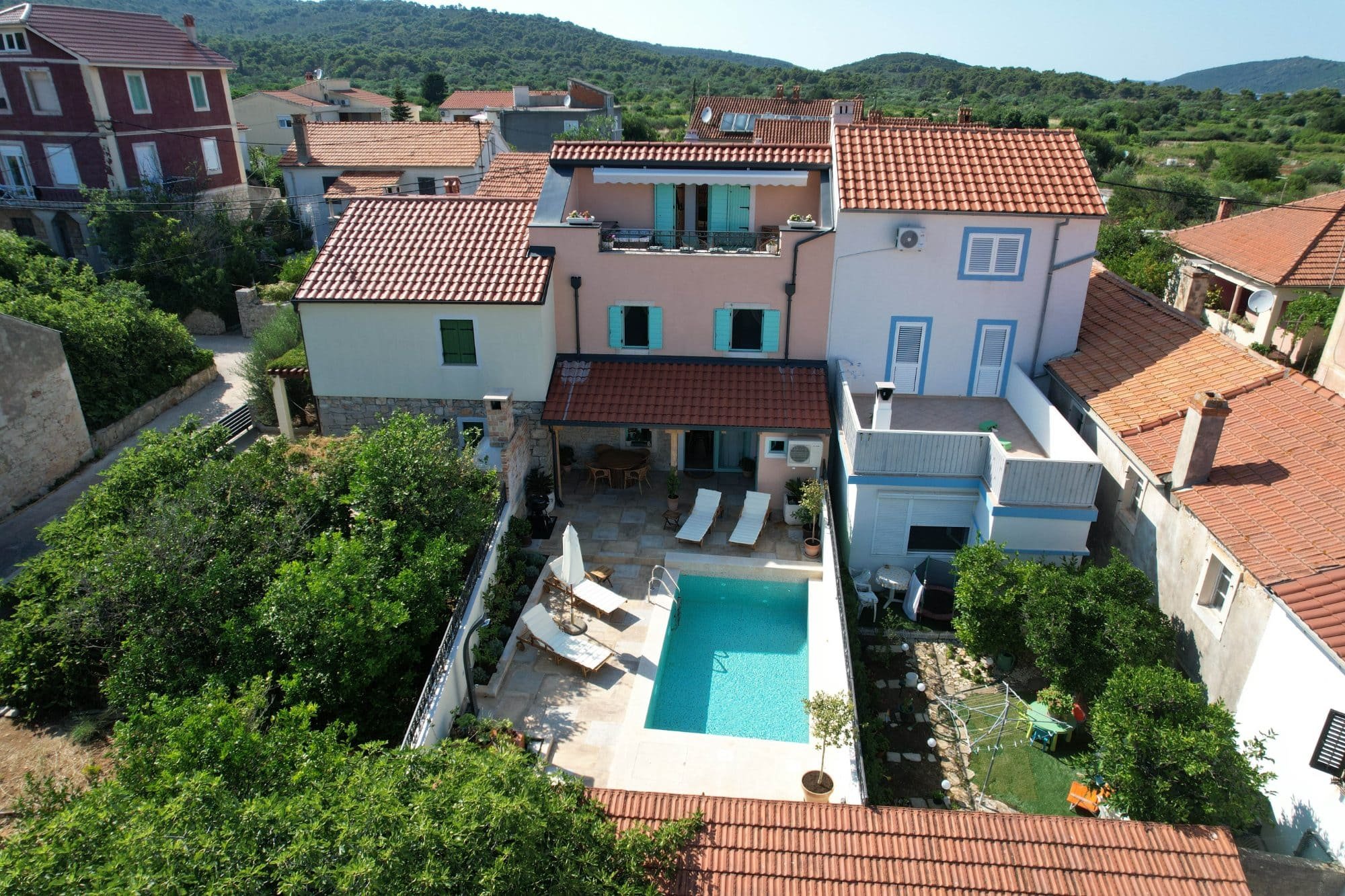 Aerial view of a residential house with a swimming pool, outdoor lounge chairs, and greenery, surrounded by other houses with red-tiled roofs in a hilly, green landscape.