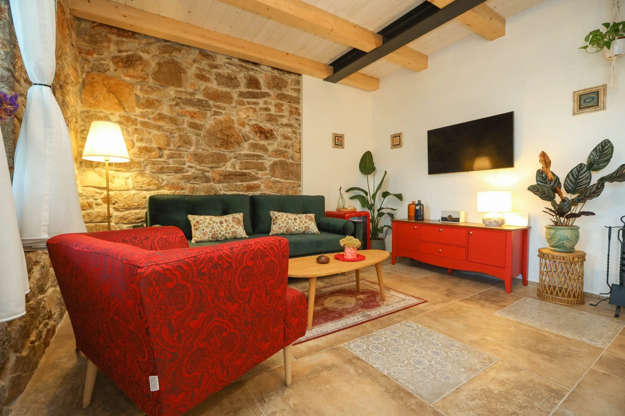 A living room with a stone accent wall, wooden ceiling beams, and various furniture and decor elements including a red patterned armchair, a green velvet sofa, a round wooden coffee table, lamps, plants, and framed artwork.