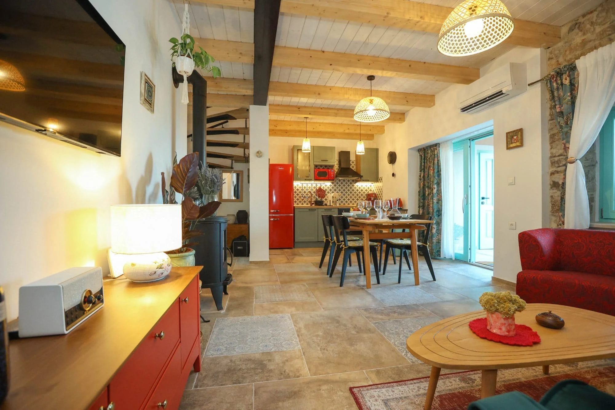 Open-concept kitchen and dining area with wooden ceiling beams, a red refrigerator, green cabinets, and a dining table with black chairs. There's a TV mounted on the wall, sliding glass door with floral curtains, and decorative items including plants