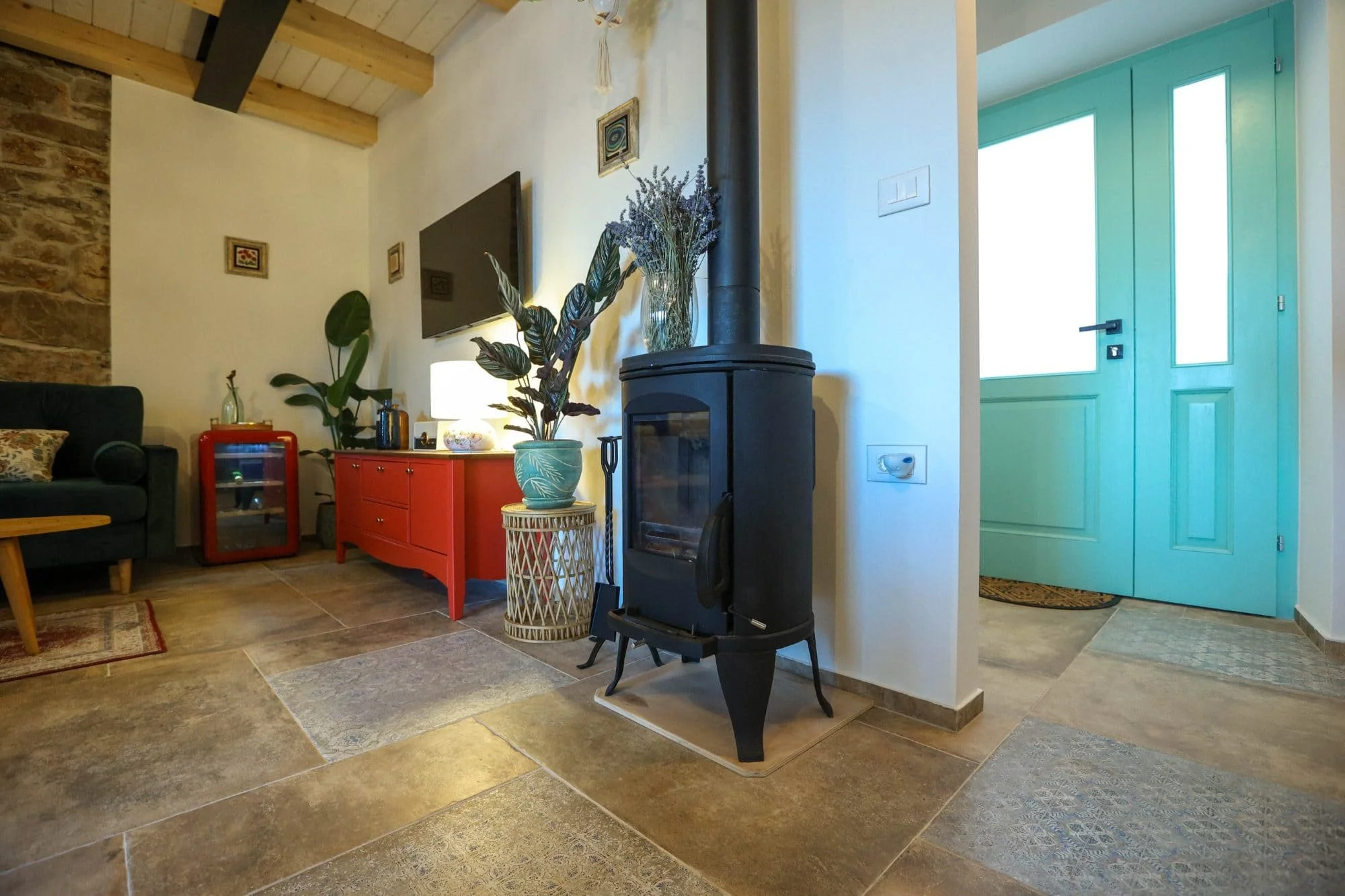 Living room with stone and white walls, a black wood stove, a green door, a red sideboard, a lamp, a potted plant, and wall art.