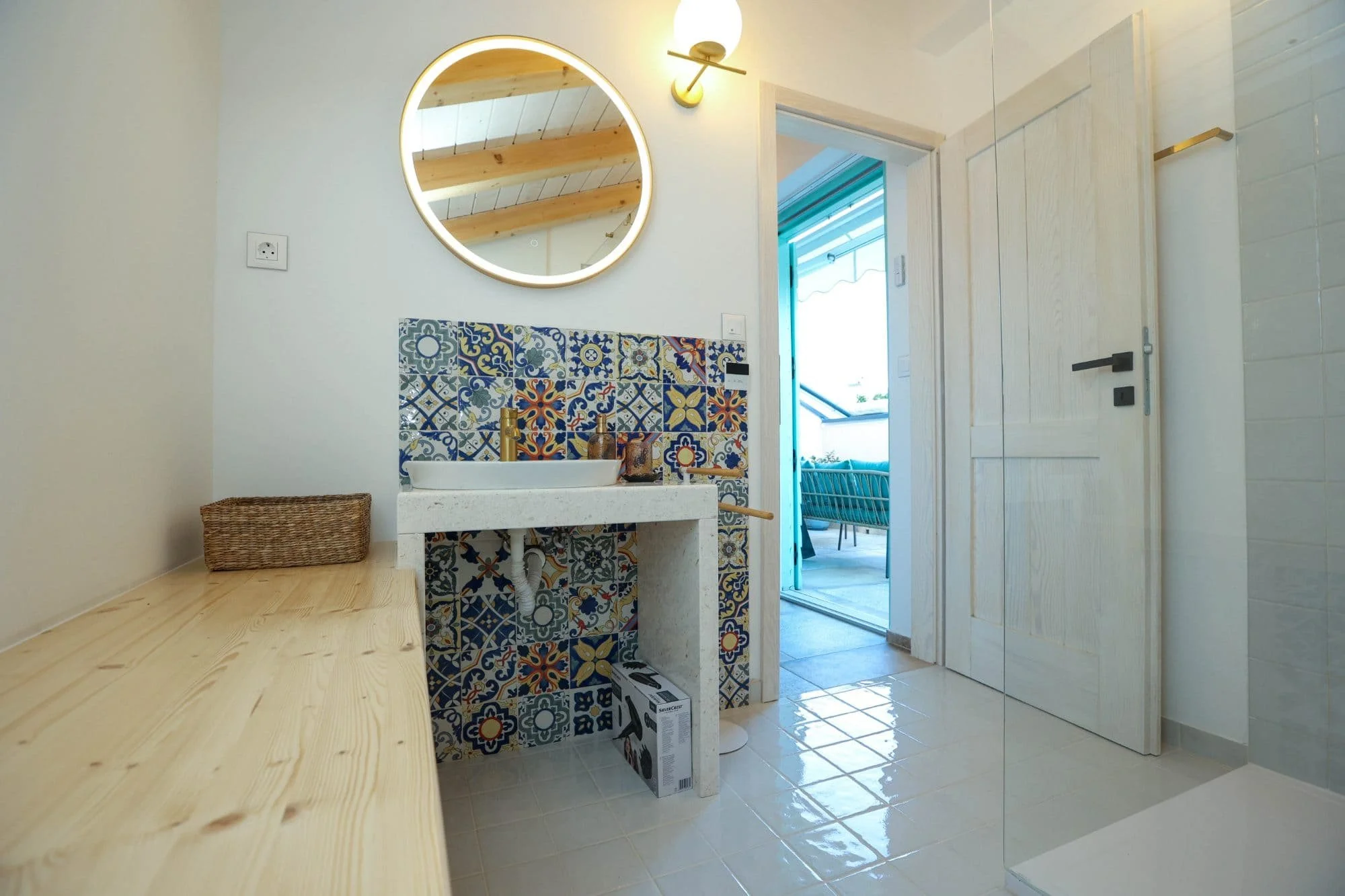 Interior of a bright, modern kitchen or bathroom area with a round mirror with backlighting, a wall-mounted light fixture, a wooden door slightly ajar, and colorful patterned tiles around a sink with a gold faucet. There is a wicker basket on a woode
