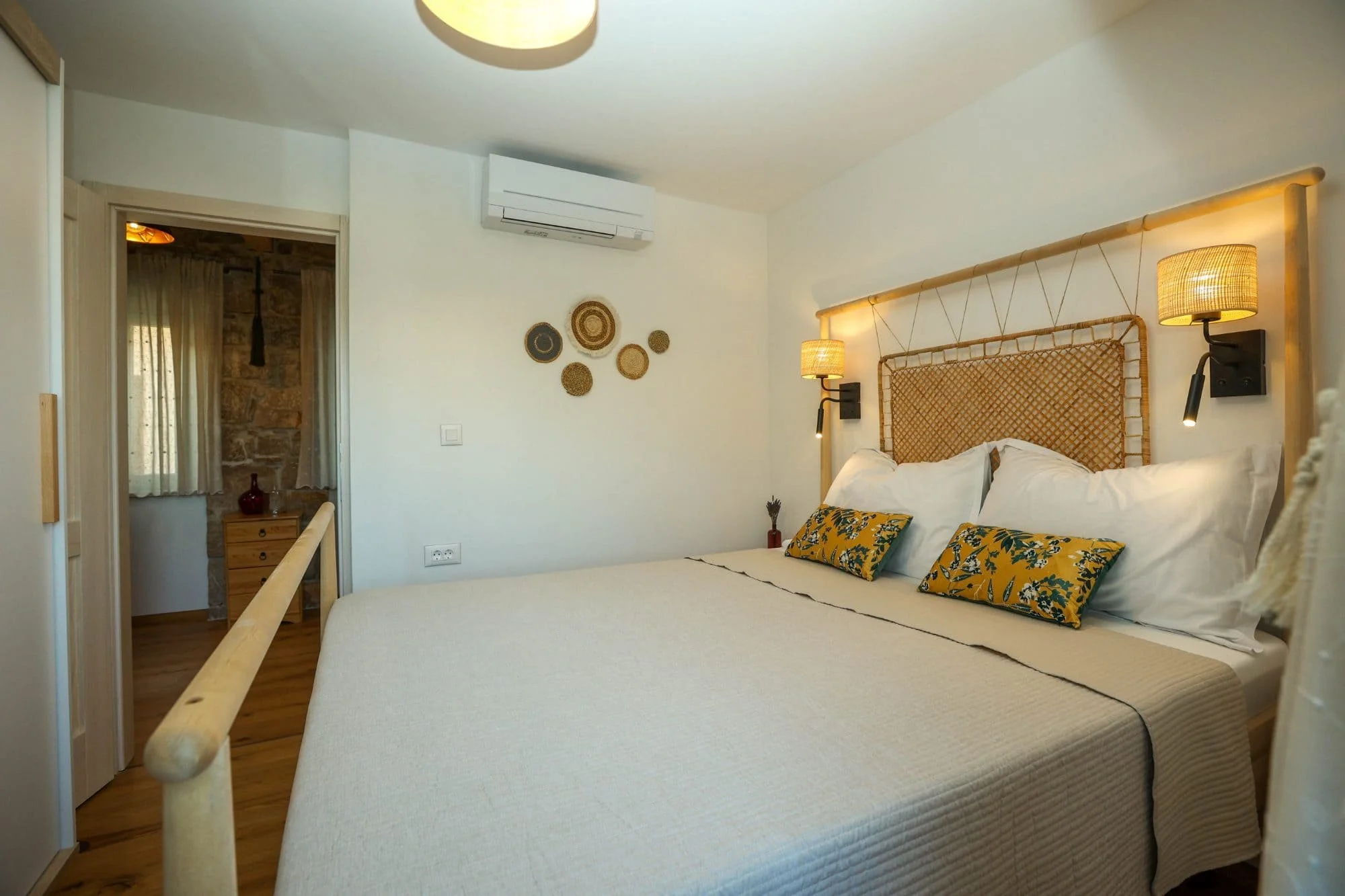 A bedroom with a bed that has decorative pillows with yellow and green floral patterns, woven rattan headboard, wall-mounted lights, and a white wall with hanging woven art. There is a doorway leading to another room with curtains and exposed brick w