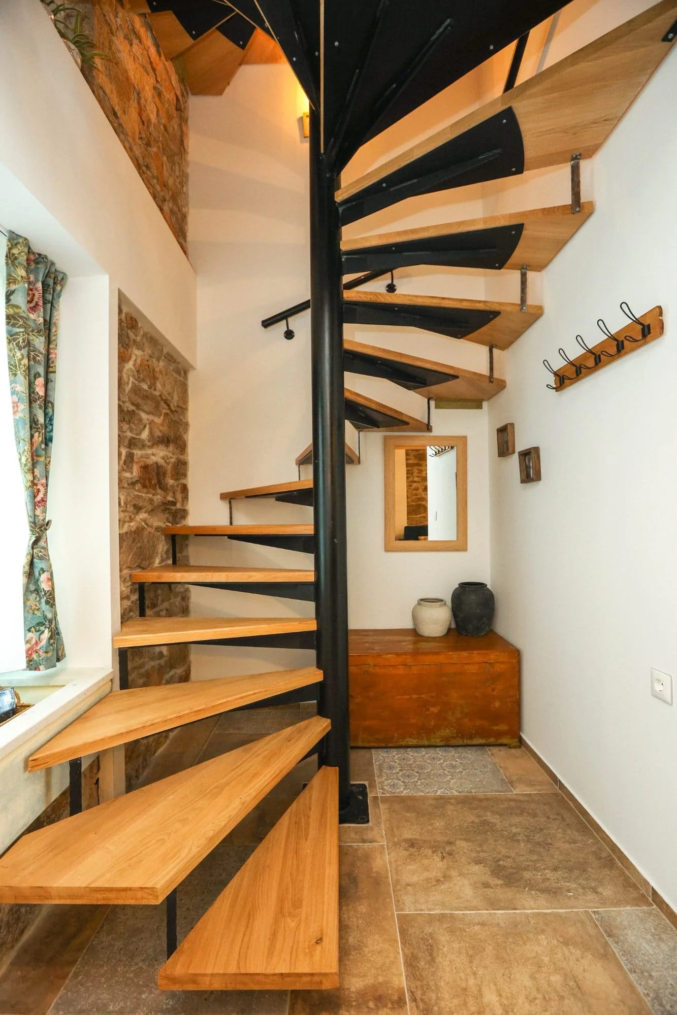 Interior view of a staircase with wooden steps and black metal supports, situated in a room with white walls, a brick accent wall, a window with floral curtains, a mirror, and decorative pots on a wooden bench.