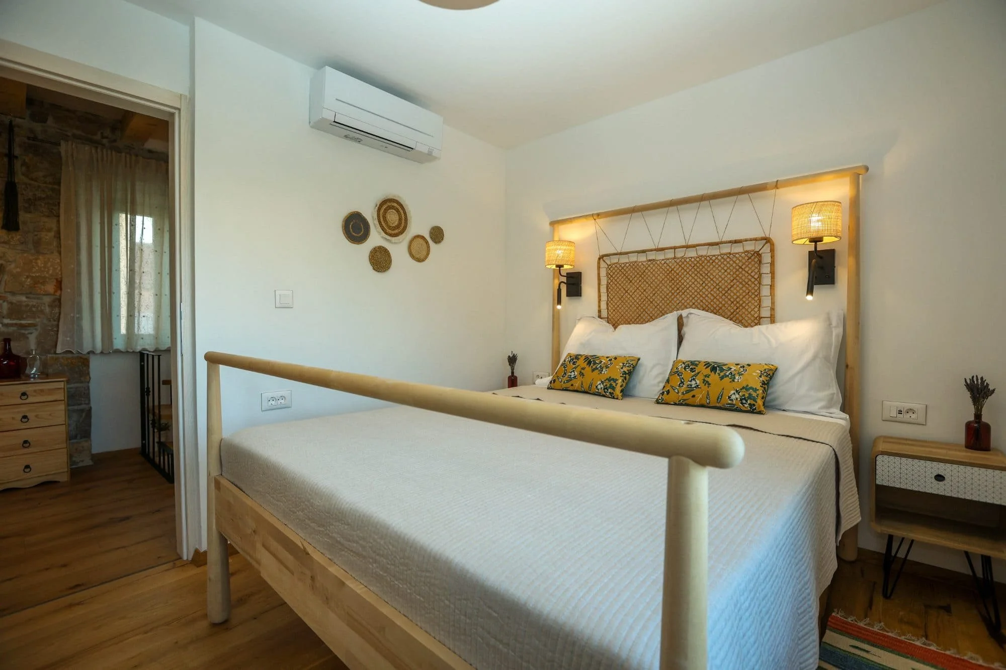 A bedroom with a wooden bed frame, white bedding, and gold-striped pillows. Two wall-mounted lamps are on either side of the bed. A small nightstand with a red vase is next to the bed. Decorative woven wall hangings are on the white wall. A wardrobe 
