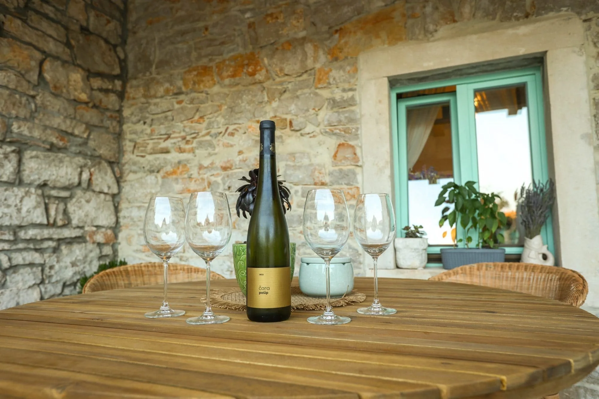 A wine bottle on a wooden table surrounded by four empty wine glasses. The background features a rustic stone wall and a window with a teal frame, showcasing potted plants and a cozy indoor setting.