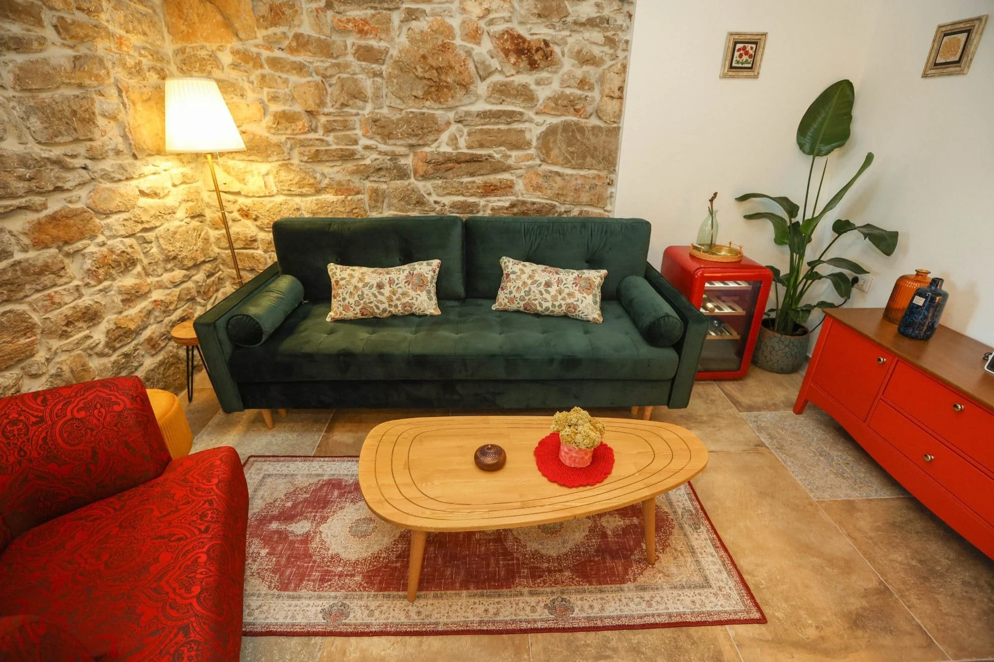 Living room with a stone accent wall, a green velvet sofa with floral pillows, a wooden coffee table with a flower arrangement, a red side table with a glass bottle, a large indoor plant in a decorative pot, framed artwork on the wall, a standing lam