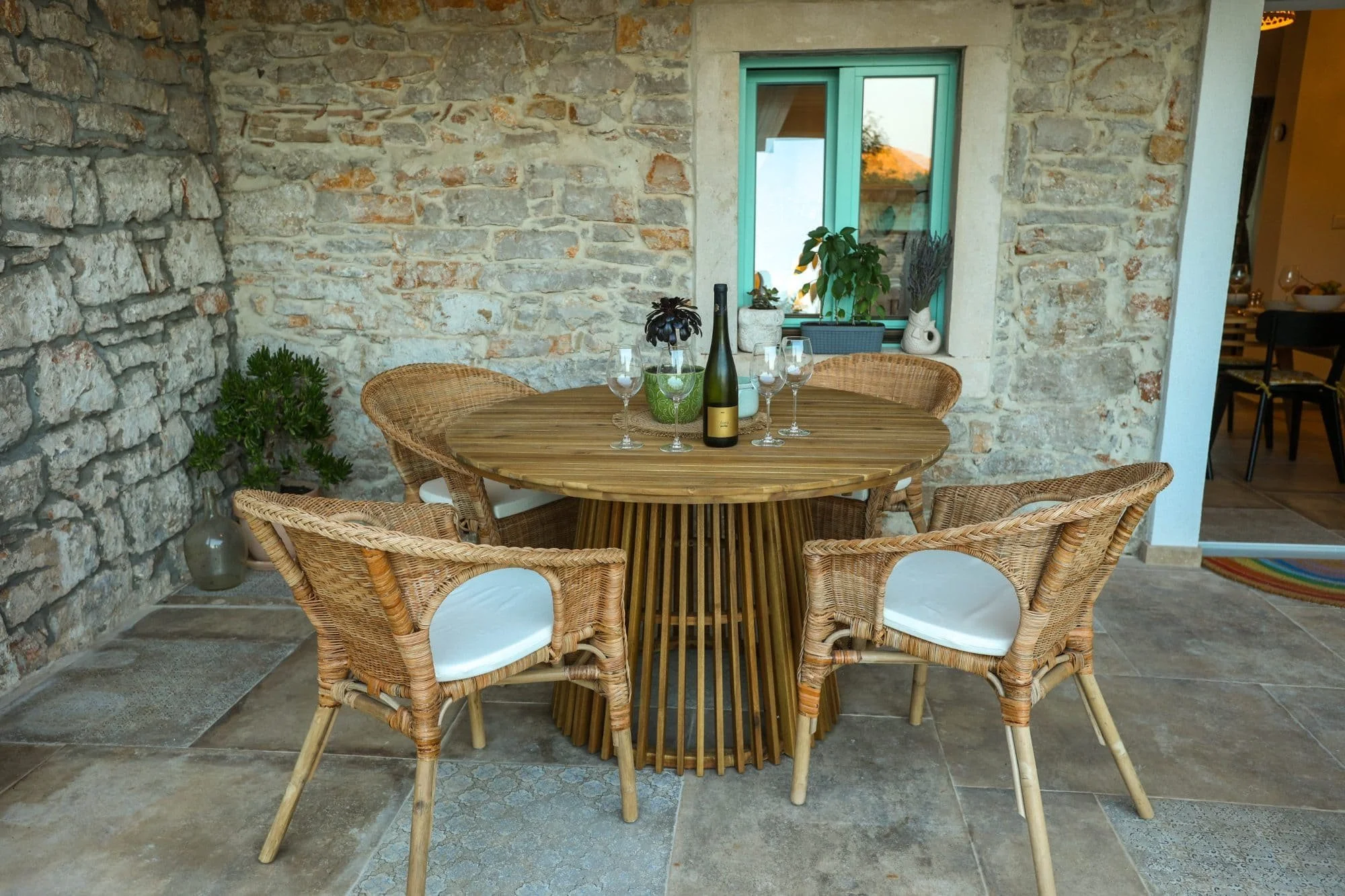 Outdoor round wooden table with six wicker chairs with white cushions, decorated with a bottle of champagne, six glasses, and potted plants, against a stone wall with a window.