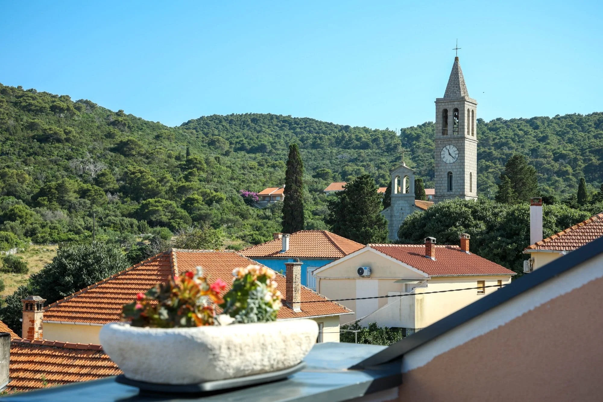 View of a town with red-tiled roofs, a church steeple, and green hills in the background under a clear blue sky. In the foreground, a balcony with a flowerpot filled with pink and white flowers.