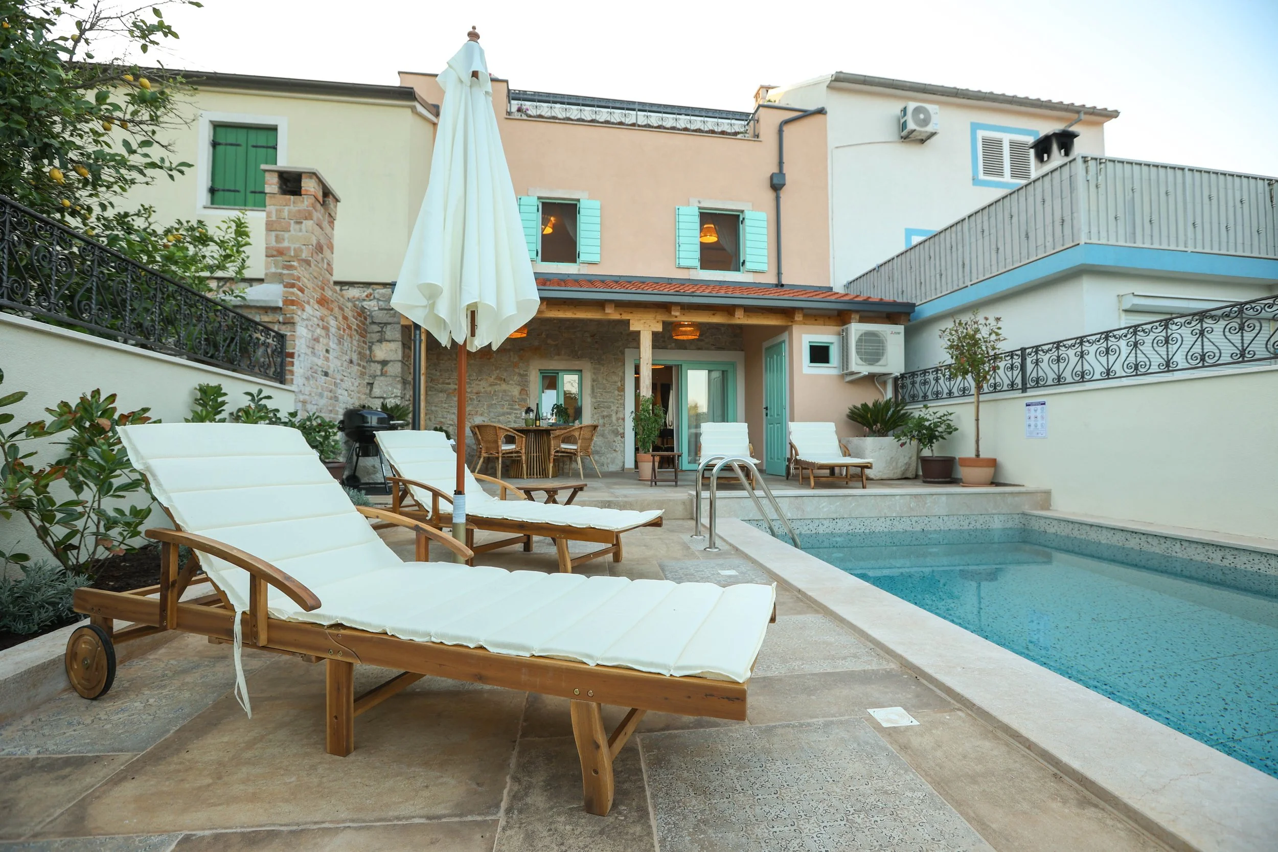 A backyard scene featuring an outdoor swimming pool, poolside lounge chairs, a patio table with chairs, a large umbrella, and a multi-story house with pastel-colored walls, green window shutters, and potted plants.