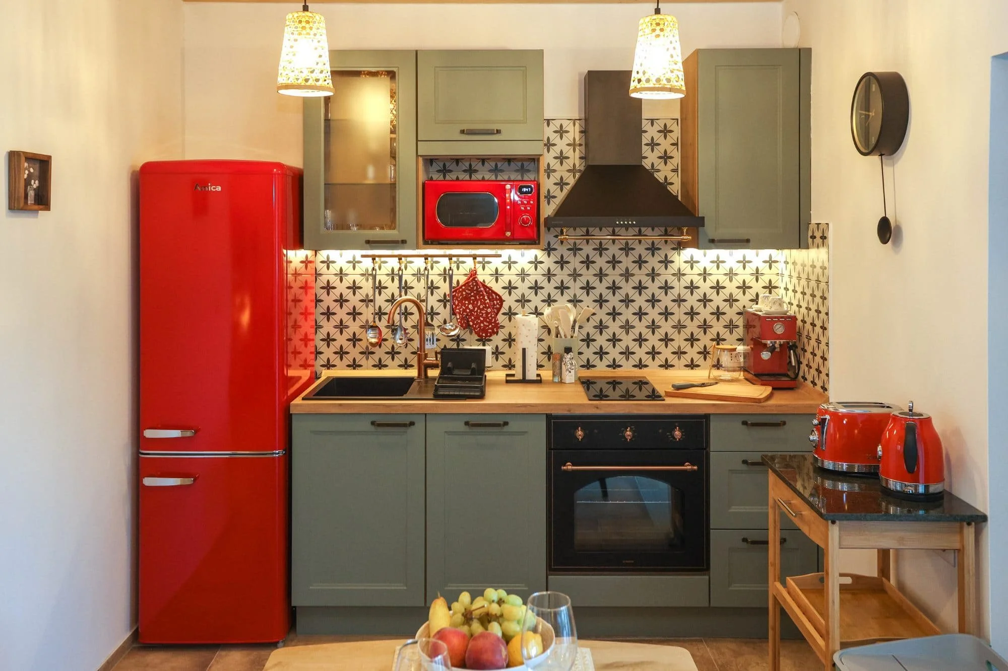 A kitchen with green cabinets, black and white patterned tile backsplash, a red refrigerator, microwave, and coffee maker, with pendant lights hanging from the ceiling.