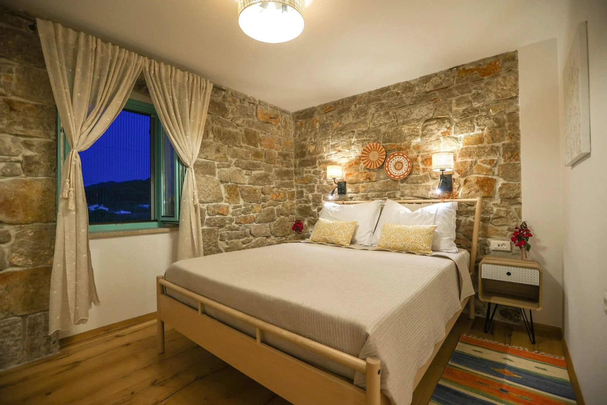 A bedroom with a stone accent wall, a bed with white bedding and yellow decorative pillows, two wall-mounted lamps on either side of the bed, and a side table with a small potted plant and a colorful striped rug. There is a window with beige curtains