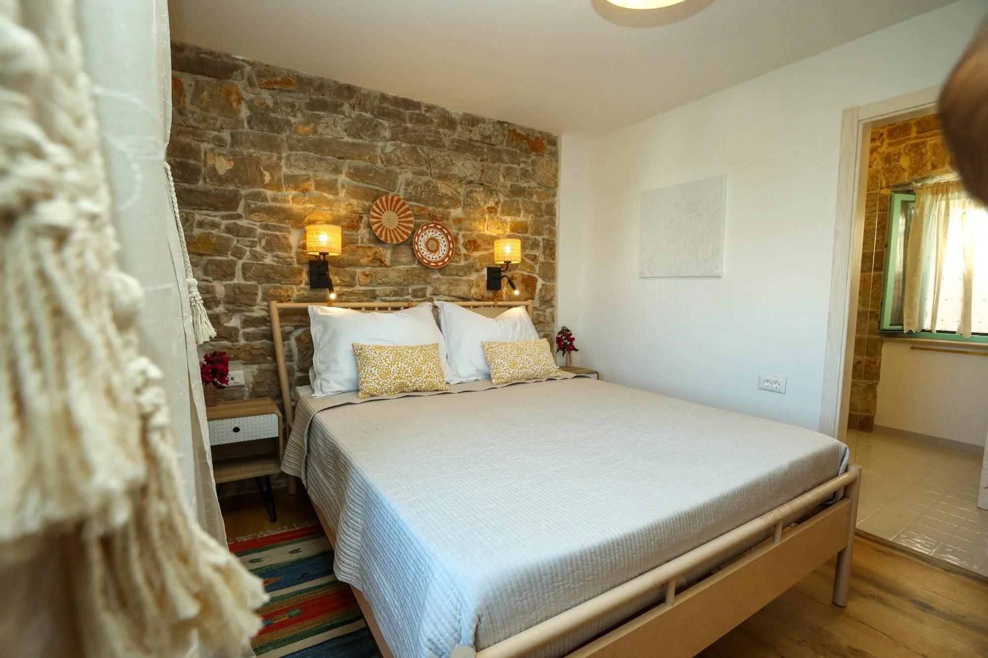 View of a cozy bedroom with an exposed brick wall, a wooden bed frame, white bedding with yellow patterned pillows, a side table with a small plant, two wall-mounted lamps, and wall decorations, with a window showing outside sunlight.