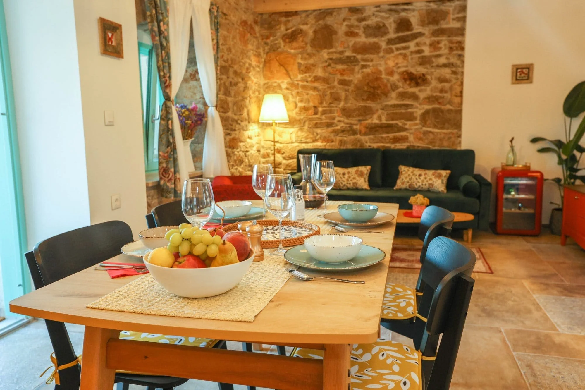 A dining table set with a fruit bowl, bowls, glasses, and cutlery in a cozy living room with exposed brick wall, a sofa with pillows, a lamp, and a window with floral curtains.