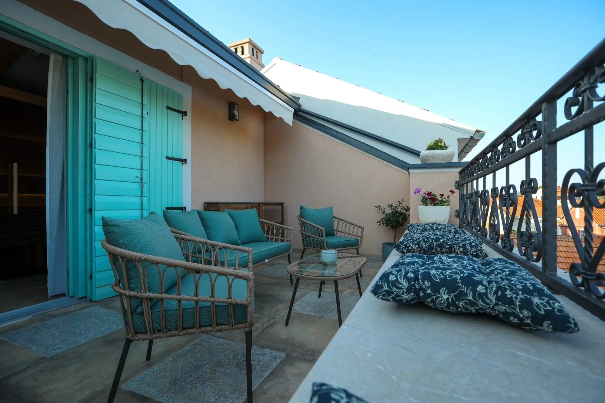 View of a rooftop terrace with outdoor furniture, including wicker chairs with teal cushions, a glass-topped table, and decorative pillows with a floral pattern. There is a small potted plant on the ledge and a blue sky overhead.