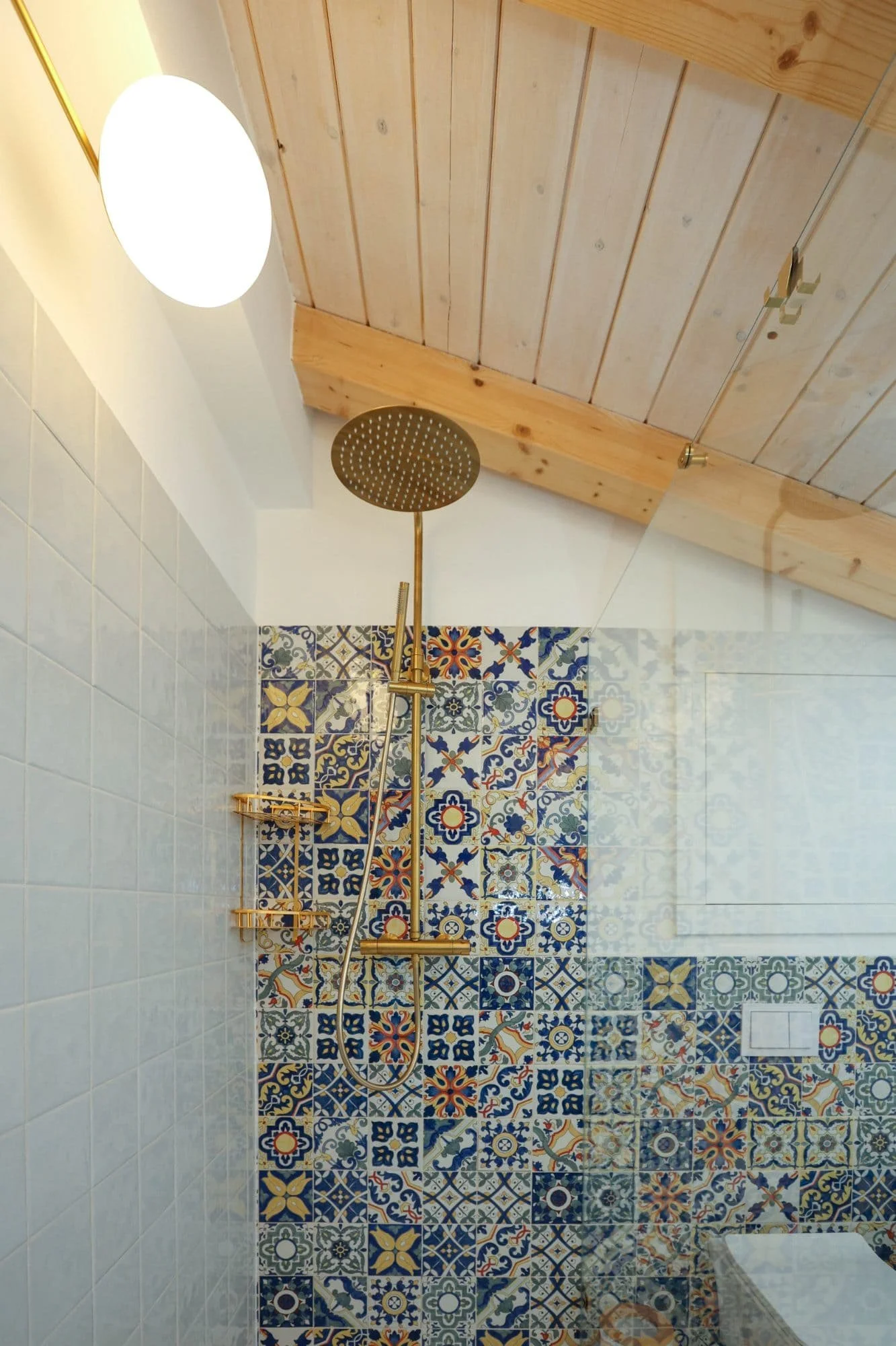 A shower area with a gold-colored showerhead and fixtures, decorated with Zellige-style patterned tiles in blue, yellow, and white. The wall has a shelf and a soap dish, with a wooden ceiling above.
