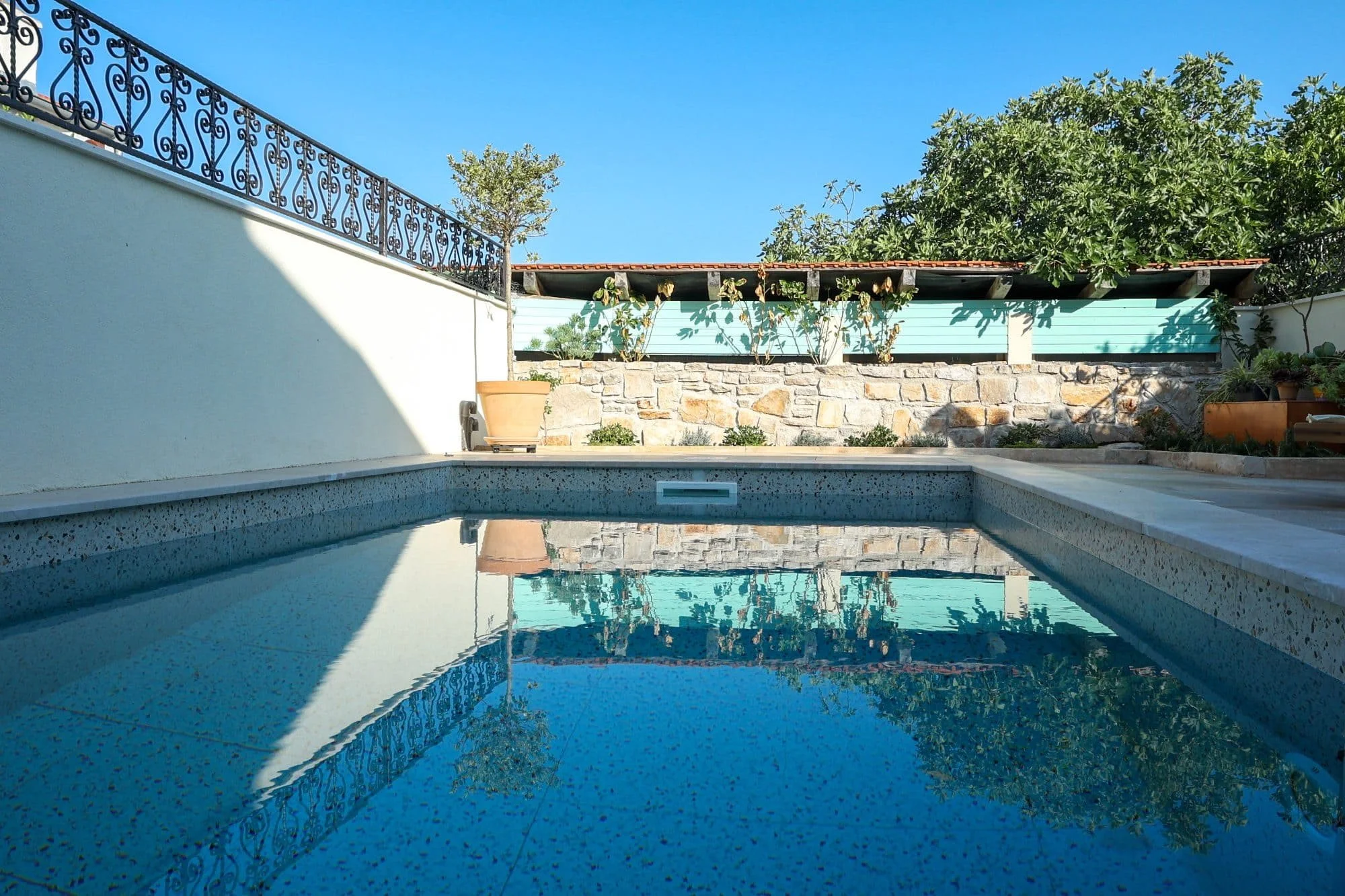 Reflecting swimming pool with blue water and a backdrop of trees, a stone wall, a porcelain pot, and a blue house wall under a clear blue sky.
