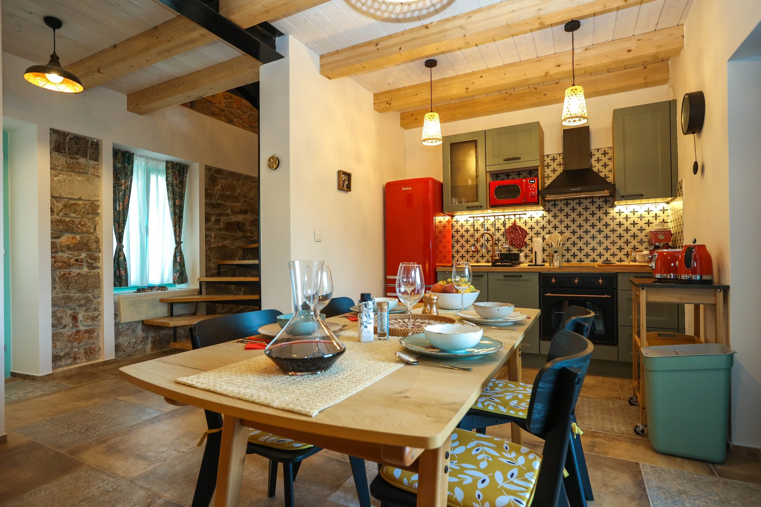 A cozy kitchen with wooden ceiling beams, hanging pendant lights, a red refrigerator, patterned backsplash, and a dining table with bowls, glasses, and chairs with yellow and white cushion covers.