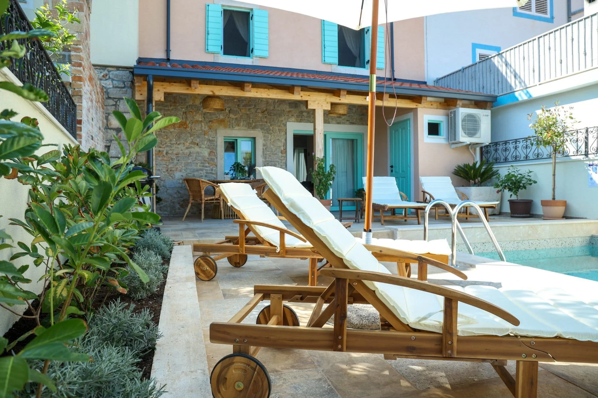 A backyard patio area with lounge chairs by a small pool, surrounded by potted plants and greenery, with a stone and stucco house featuring blue shutters and doors in the background.