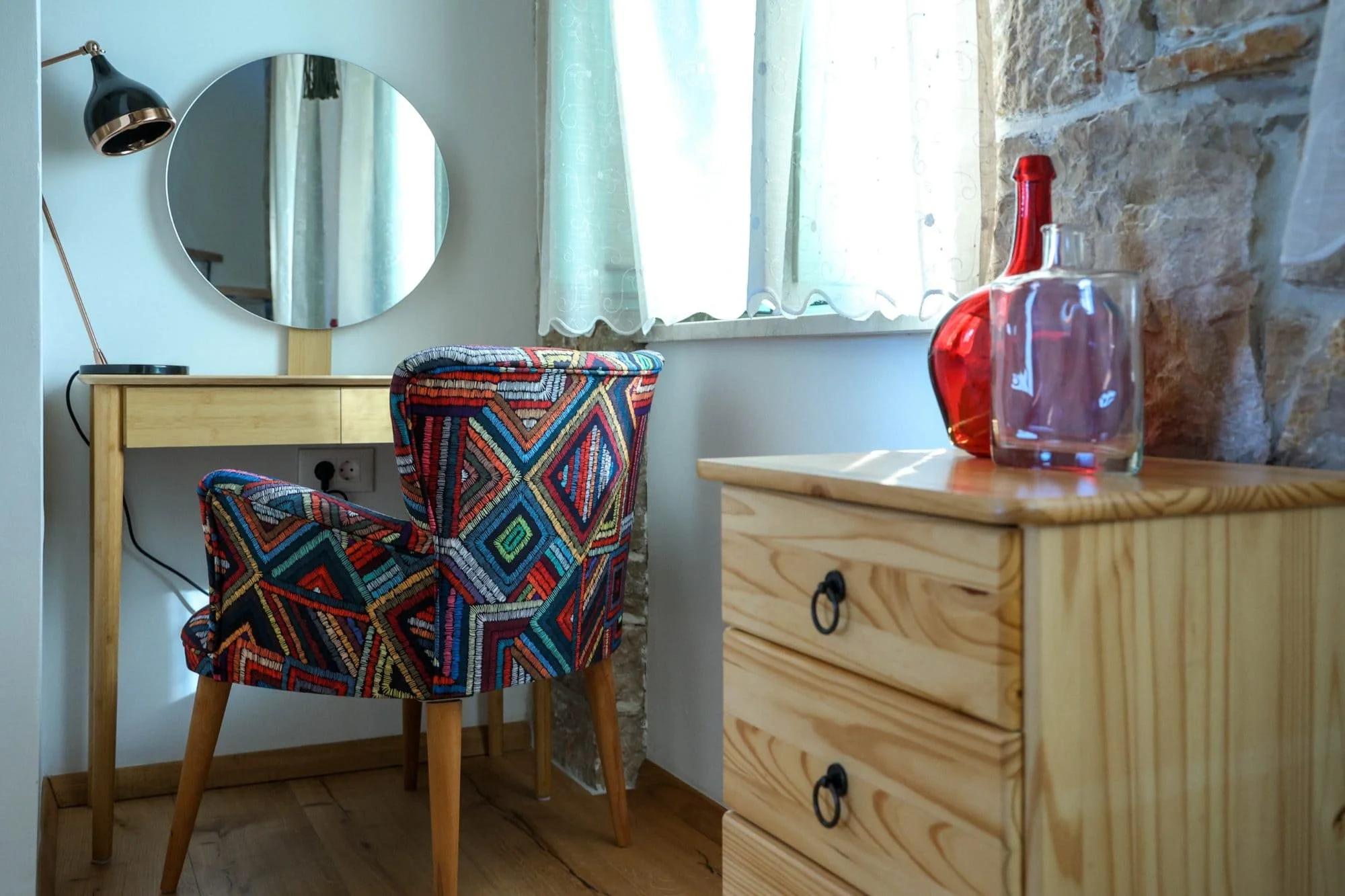 A cozy corner with a wooden desk, a patterned upholstered chair, a mirror, a desk lamp, and a wooden chest of drawers next to a window with light-colored curtains, decorated with glass vases.