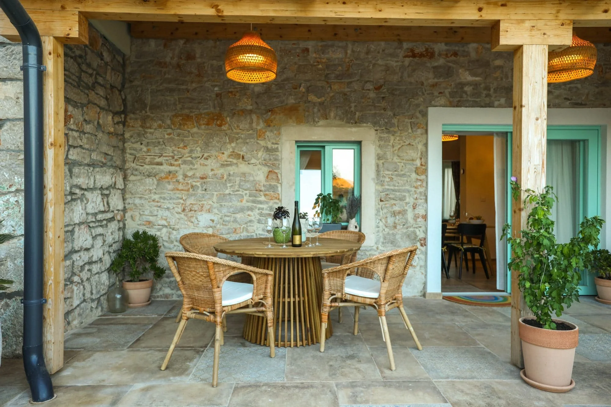 Outdoor patio area with a round dining table and wicker chairs set against a stone wall with a window and door, decorated with potted plants and hanging wicker lamps.