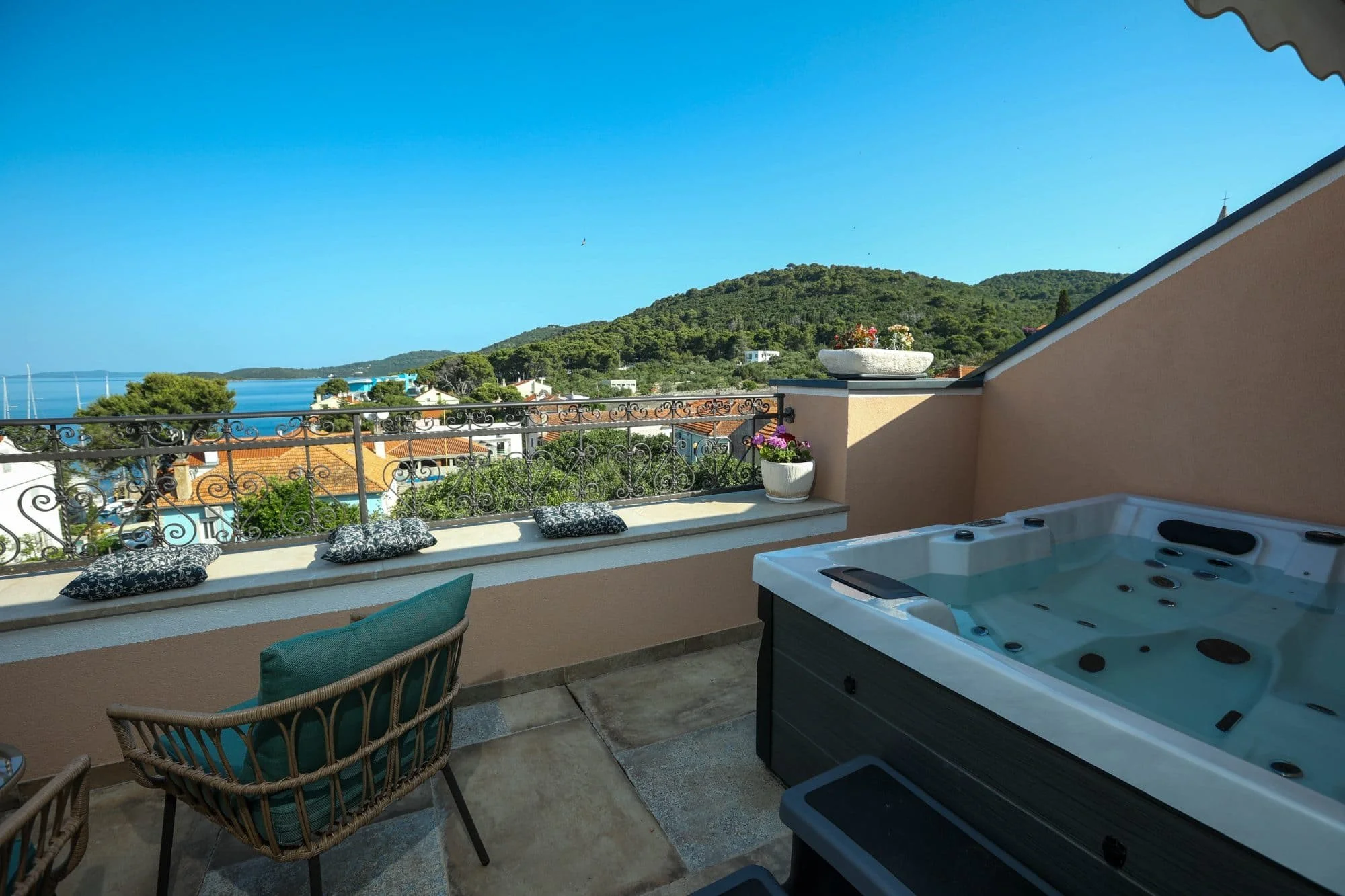 Balcony with hot tub, outdoor chairs, decorative pillows, potted plants, scenic view of hills, water, and sailboats under clear blue sky.
