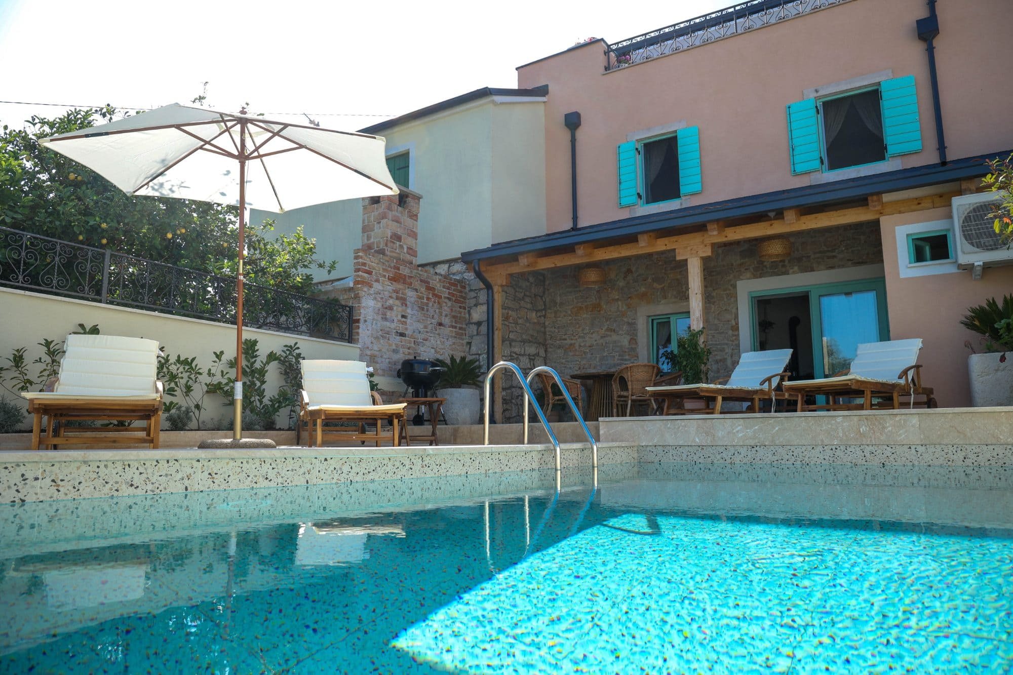 A backyard with a swimming pool, poolside chairs, a patio area with tables and chairs, an umbrella, and the back of a house with blue shutters.