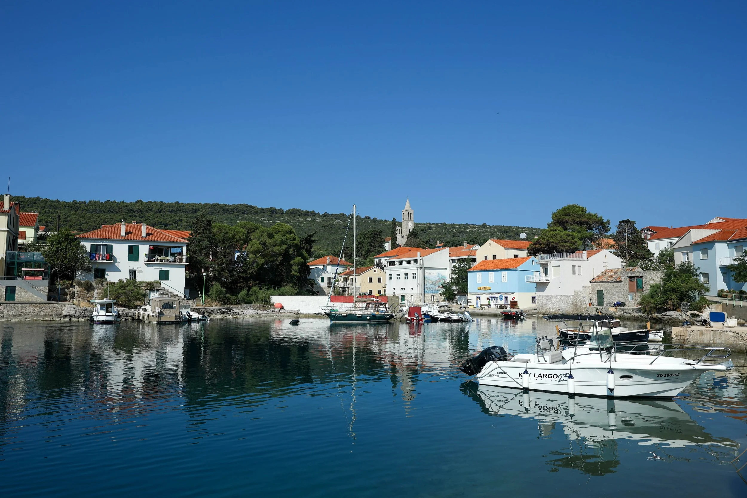 View of a small harbor with boats docked by colorful houses, a church tower on a hill, and a bright blue sky.