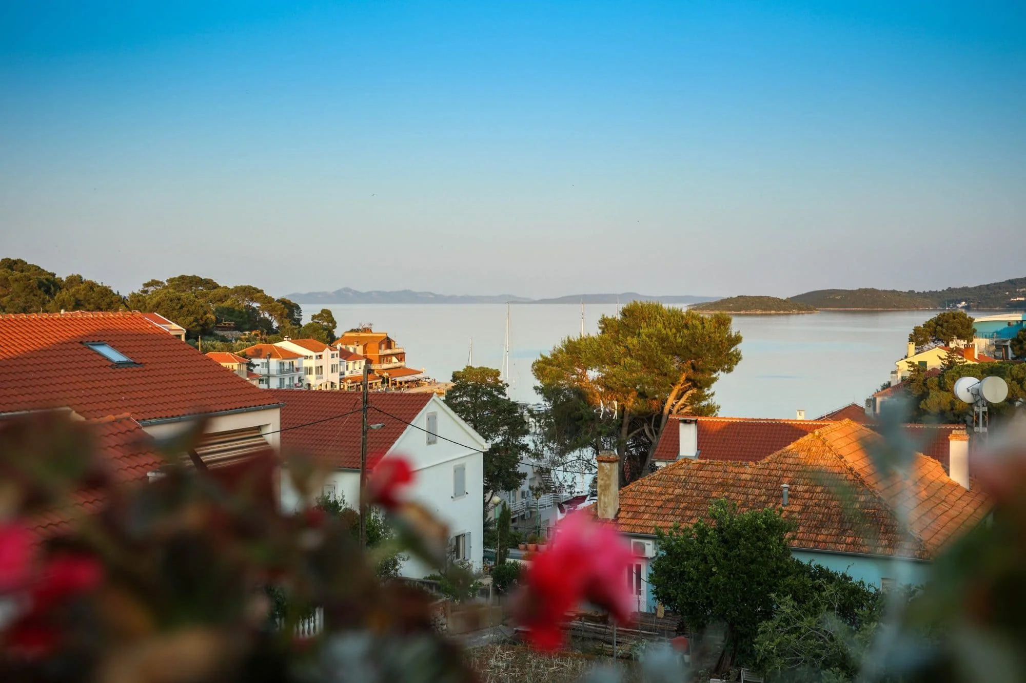 A coastal neighborhood with red-tiled roofs, trees, and boats on a calm body of water under a clear blue sky.