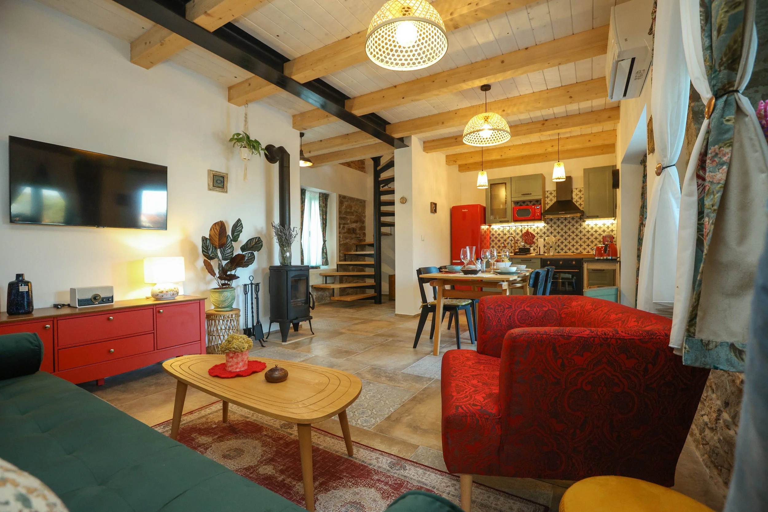 A cozy living room with wooden beams on the ceiling, a wall-mounted flat-screen TV, a red sideboard, a green sofa, a red patterned armchair, and a wooden coffee table with decorative items, with a view into a kitchen area with a red refrigerator, patterned backsplash, and dining table.
