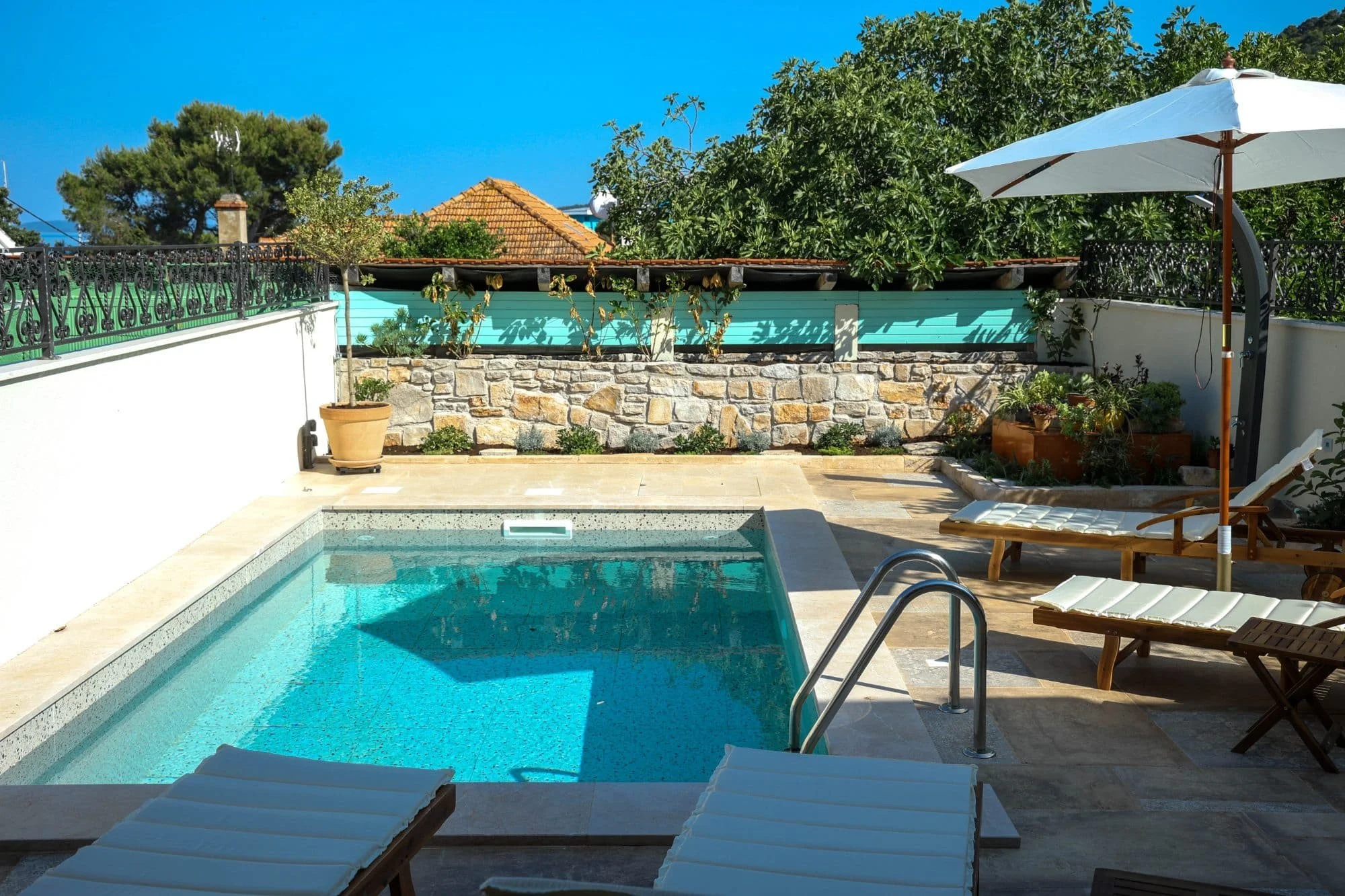 Swimming pool with lounge chairs and an umbrella in a backyard, surrounded by greenery and a stone wall under a bright blue sky.